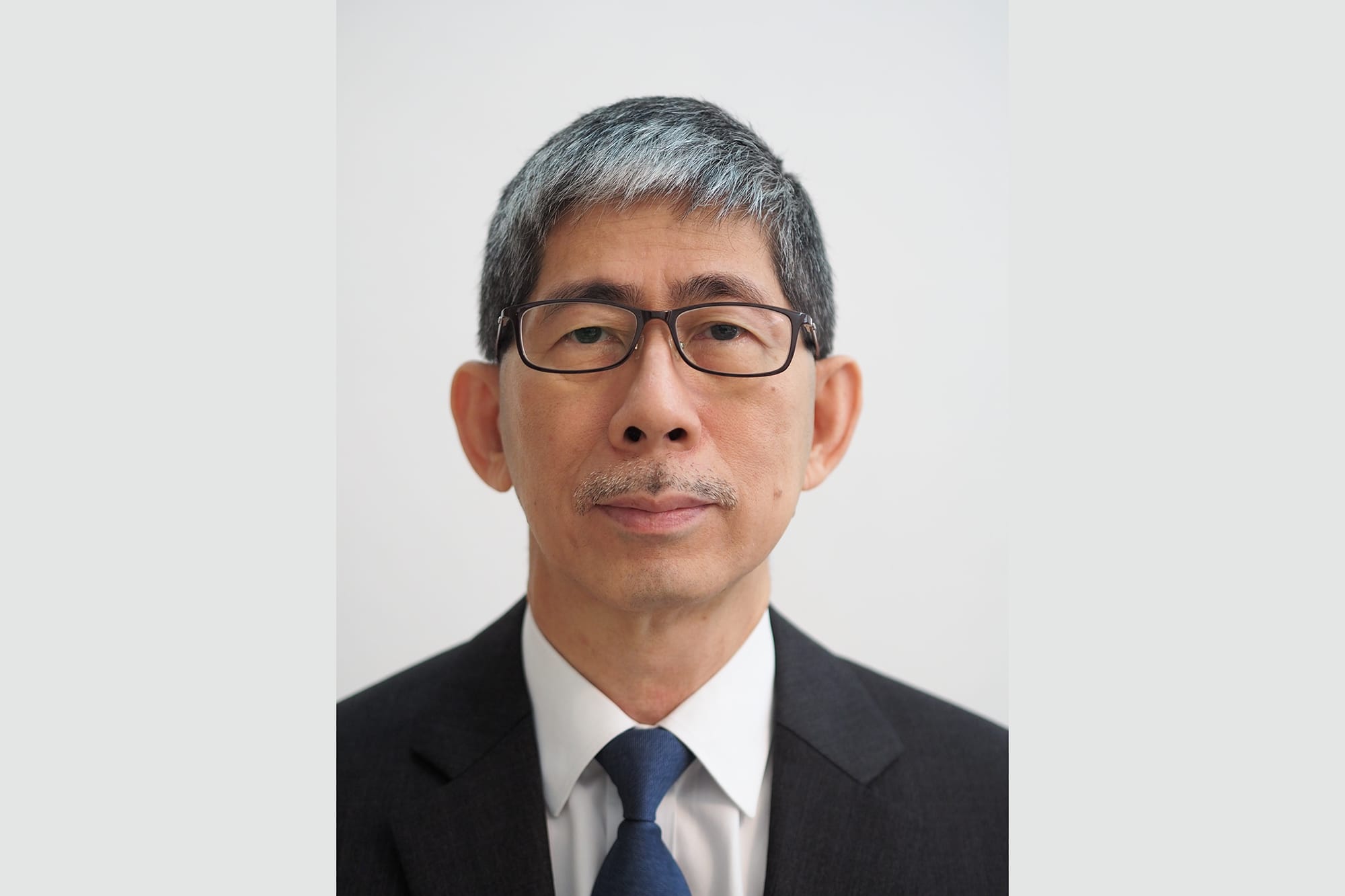 Headshot of an Asian man with gray hair and glasses in a suit and blue tie.