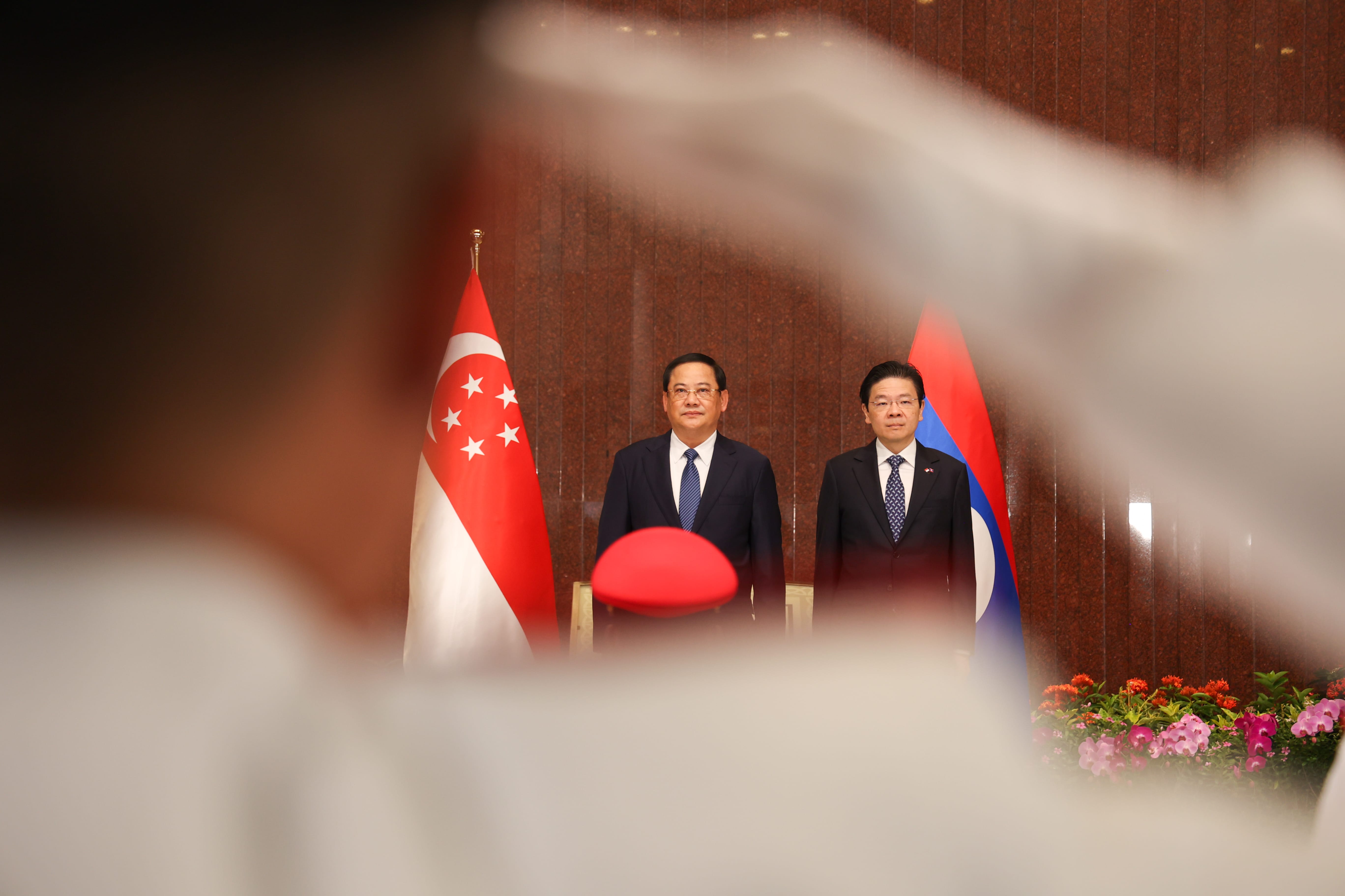 Two men in suits stand before Singapore and Laos flags; blurry foreground.