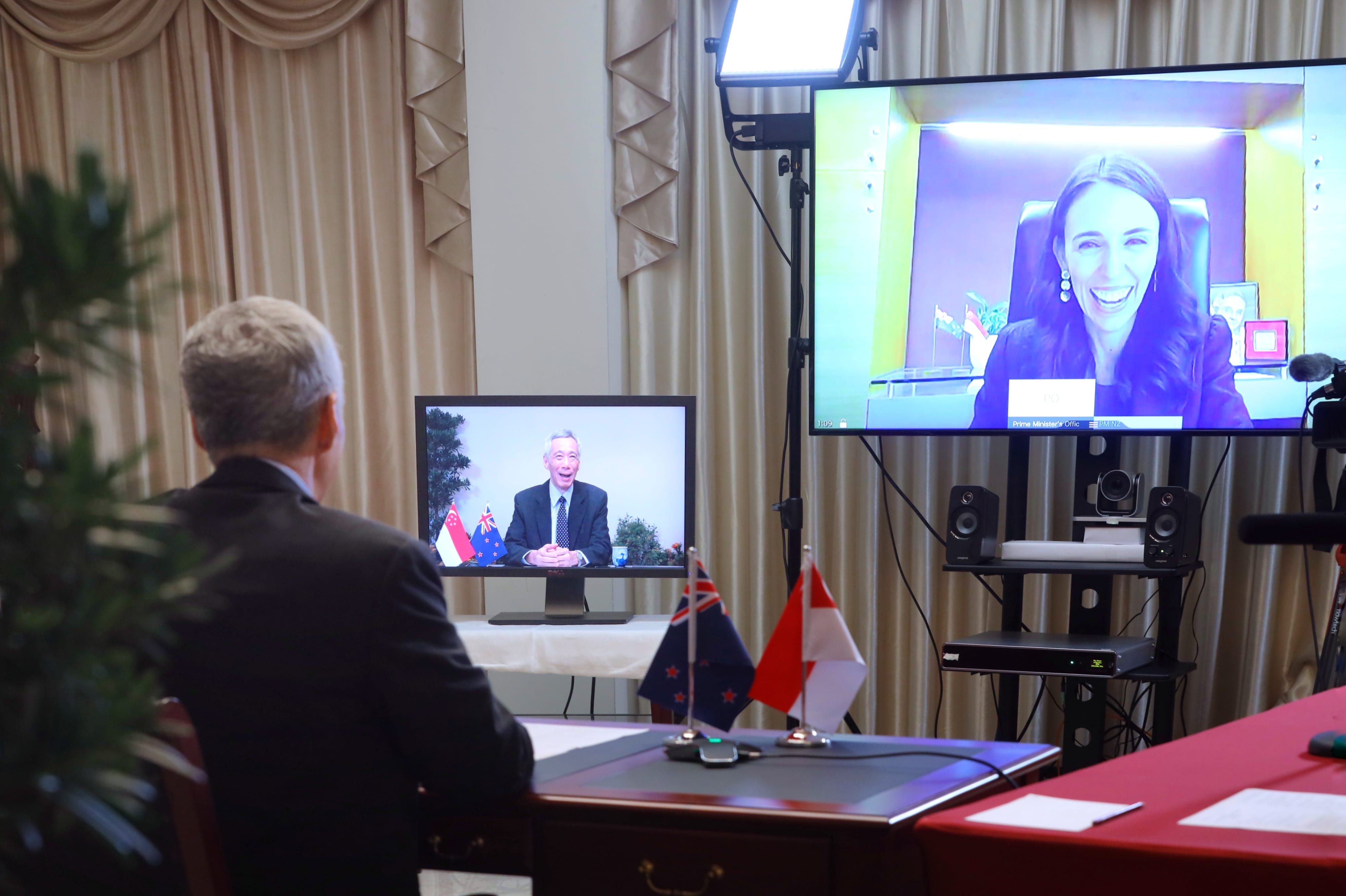 Man in suit at desk, video screens showing Jacinda Ardern and another man, New Zealand/Singapore flags.