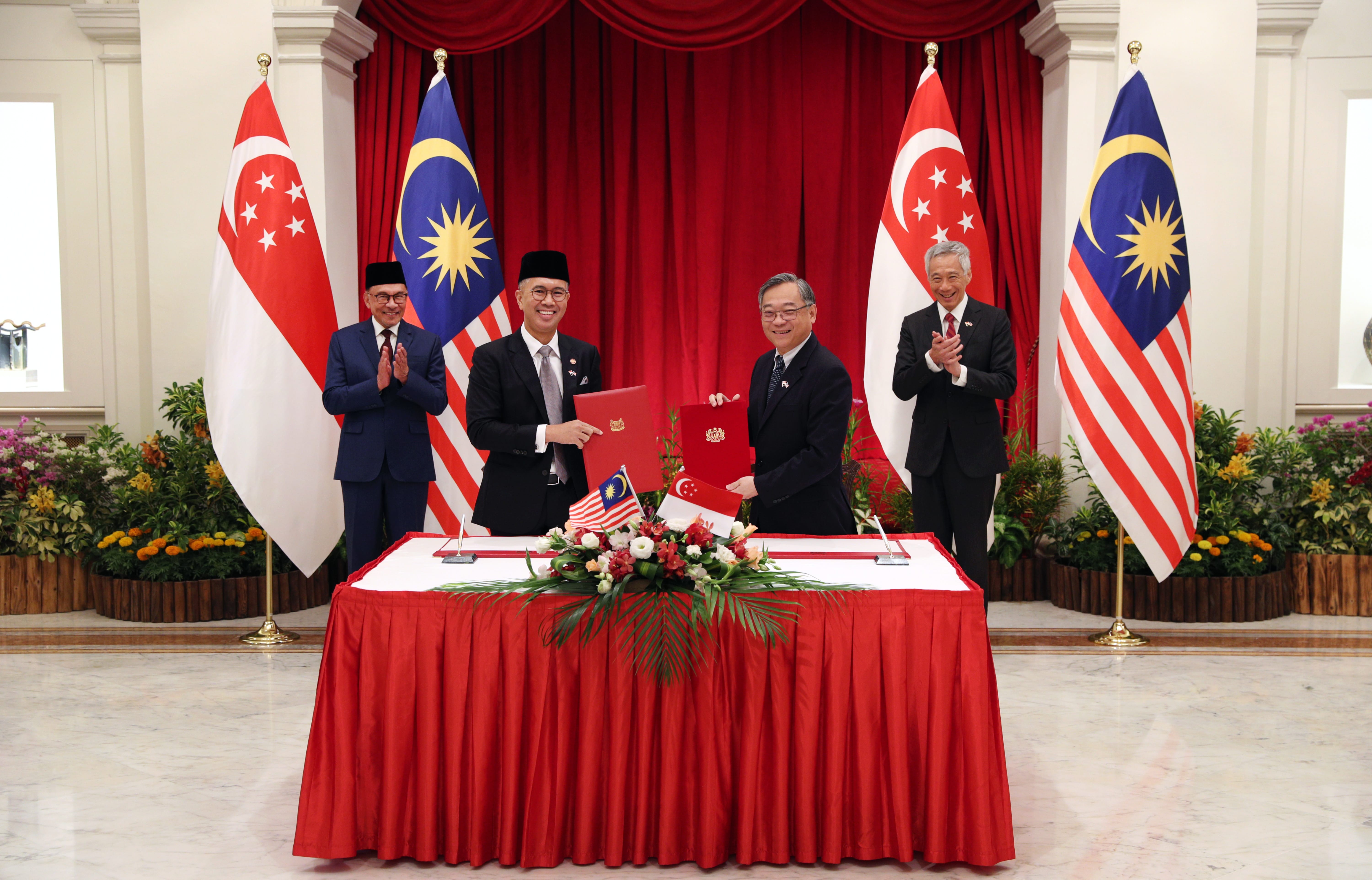 Two men in suits sign documents; flags of Singapore and Malaysia in background.