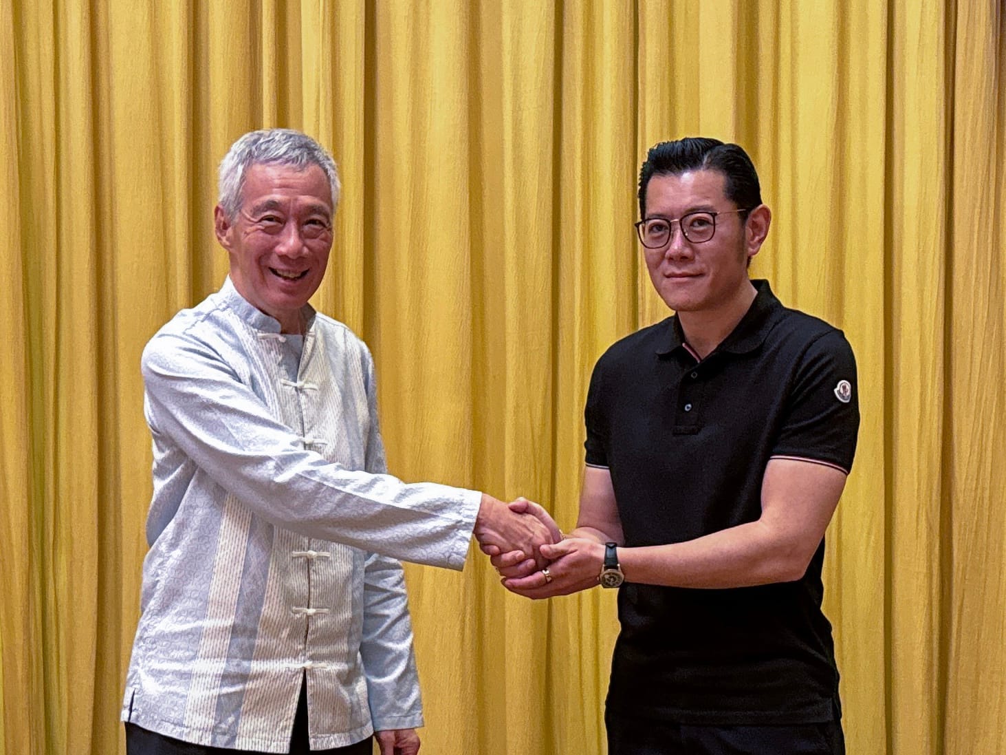 Lee Hsien Loong shaking hands with a man in black Moncler polo in front of a gold curtain.