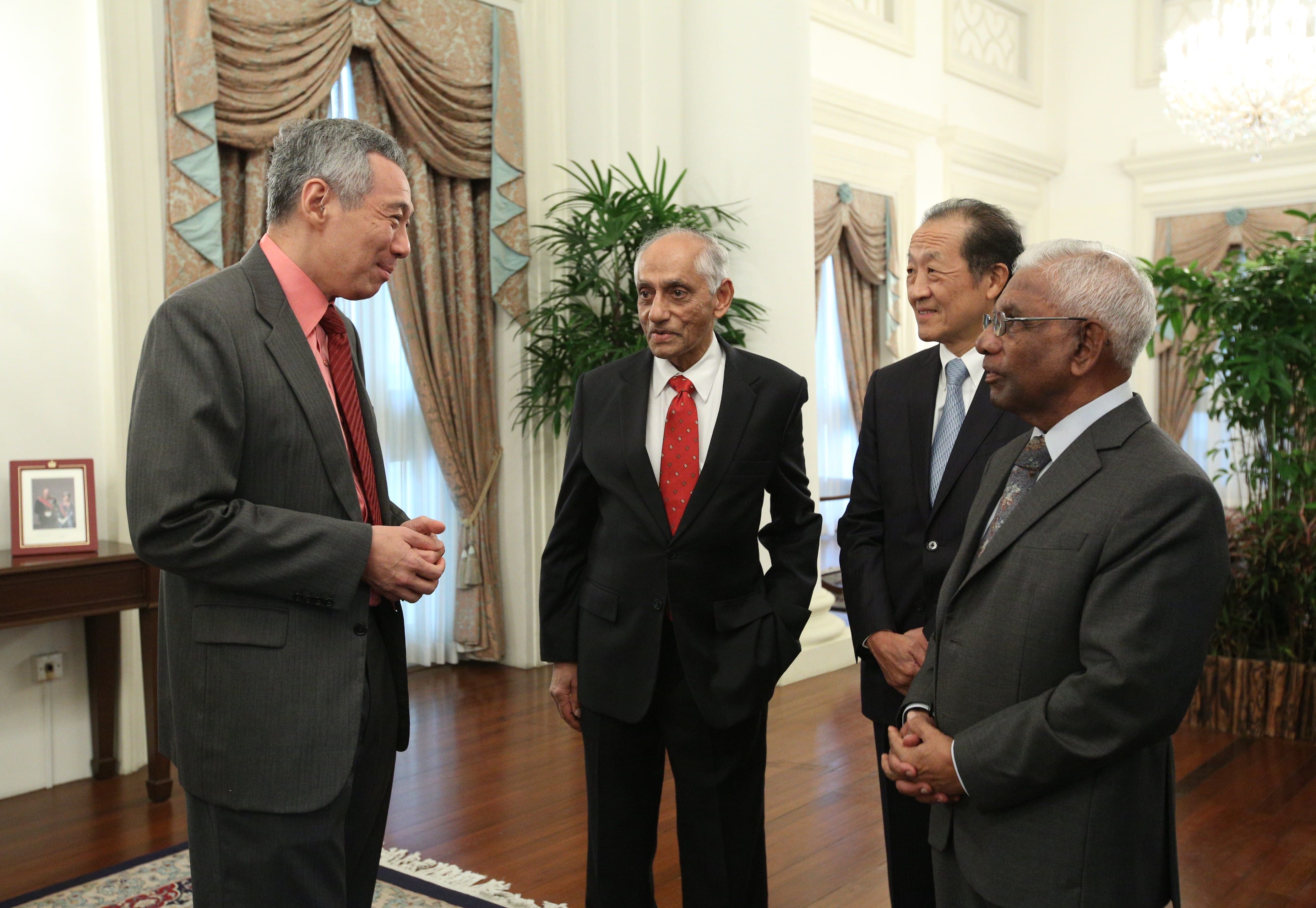 Four men in suits conversing in an ornate room with plants and a chandelier.