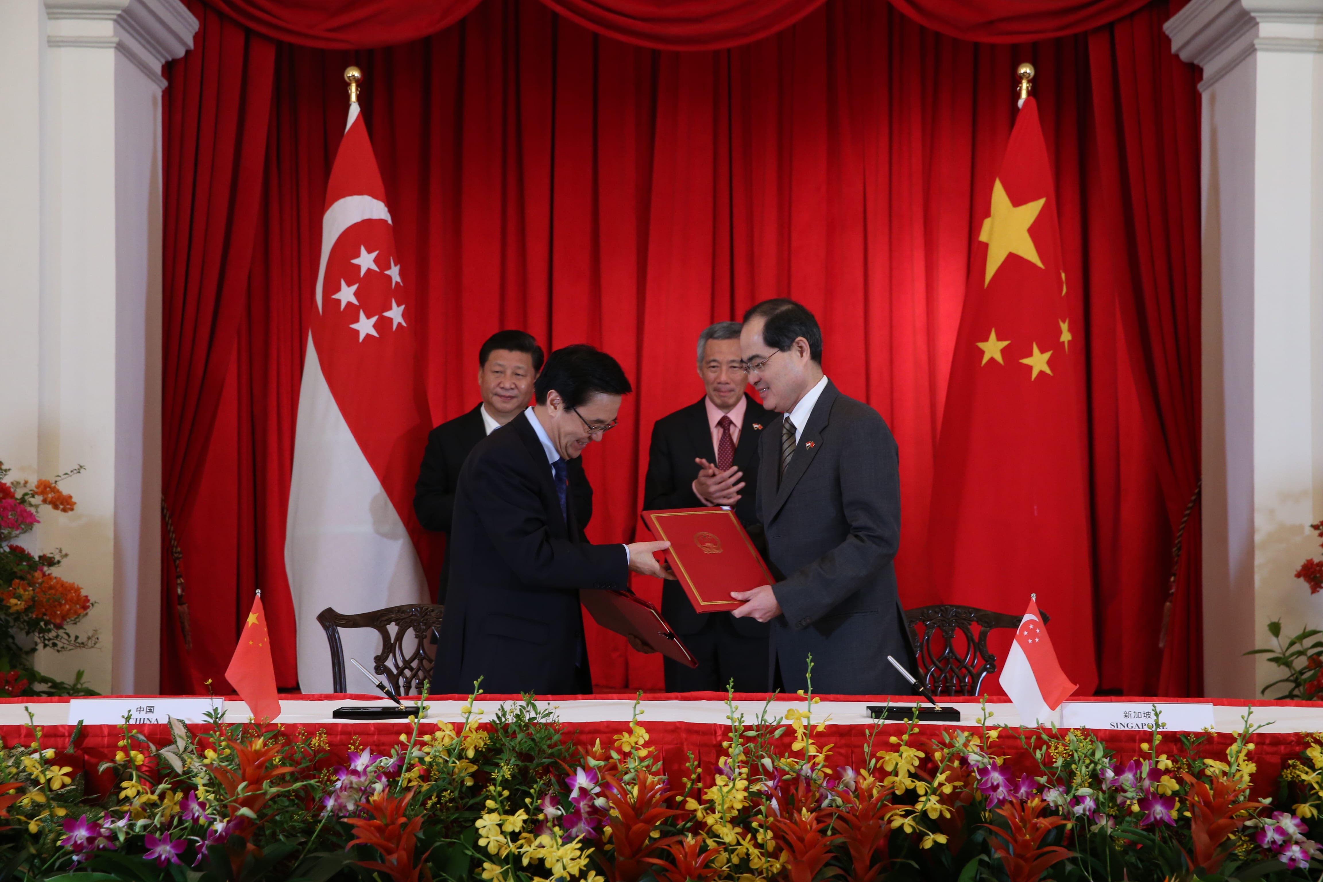 Two officials exchanging documents with flags of China and Singapore behind them.