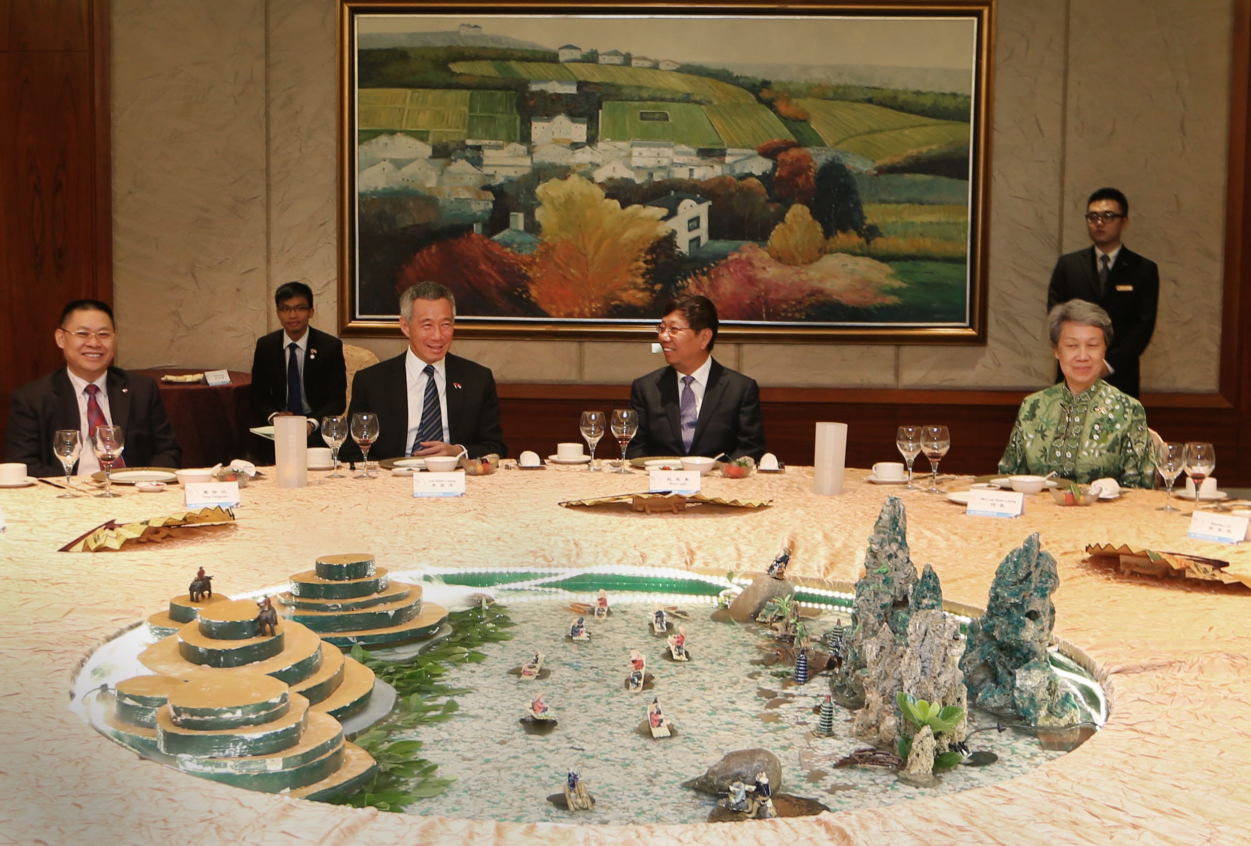People at a round table with a diorama-style centerpiece, an ornate landscape painting behind.