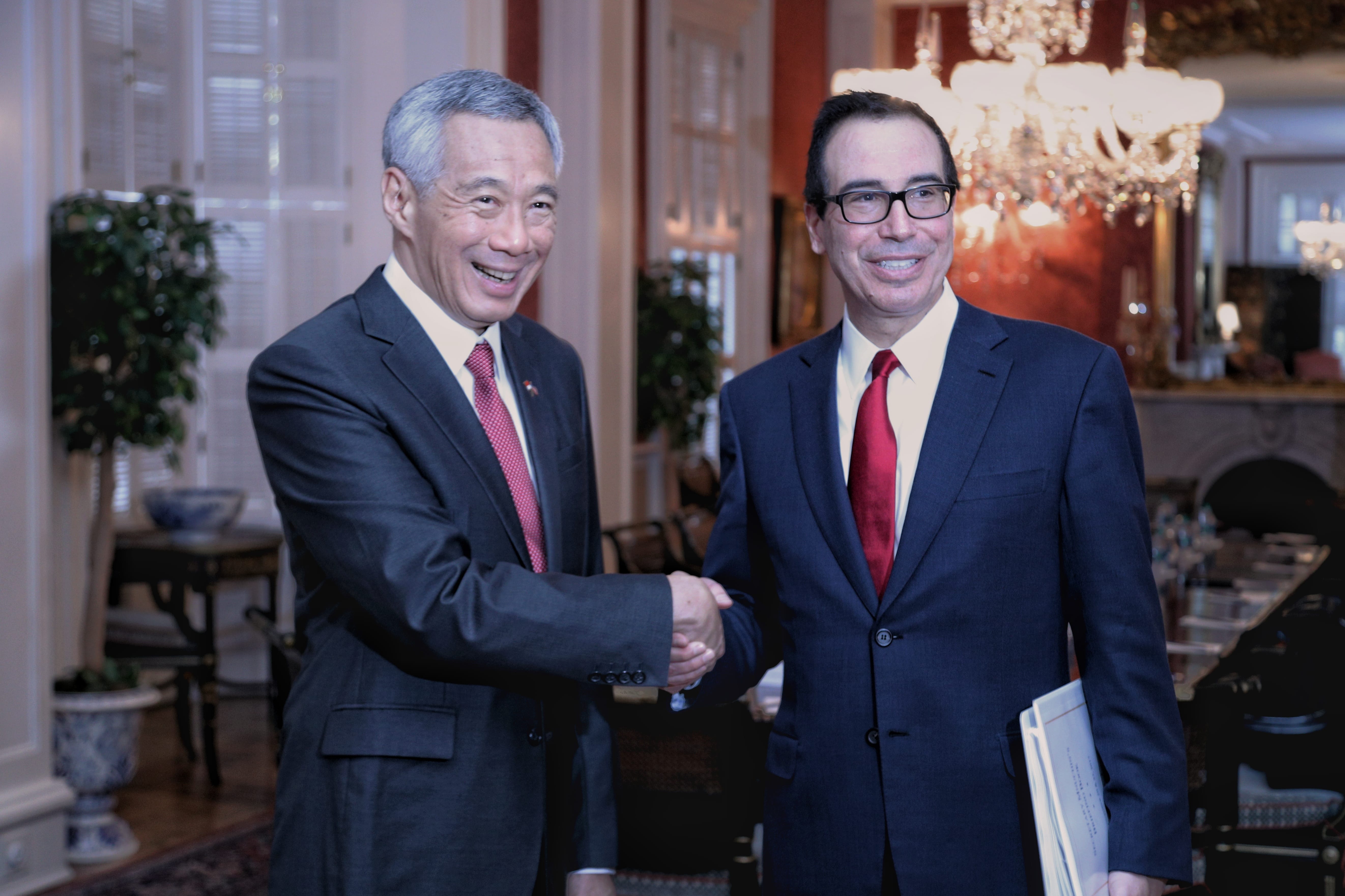 Lee Hsien Loong and Steven Mnuchin in suits shake hands in a room with a chandelier.