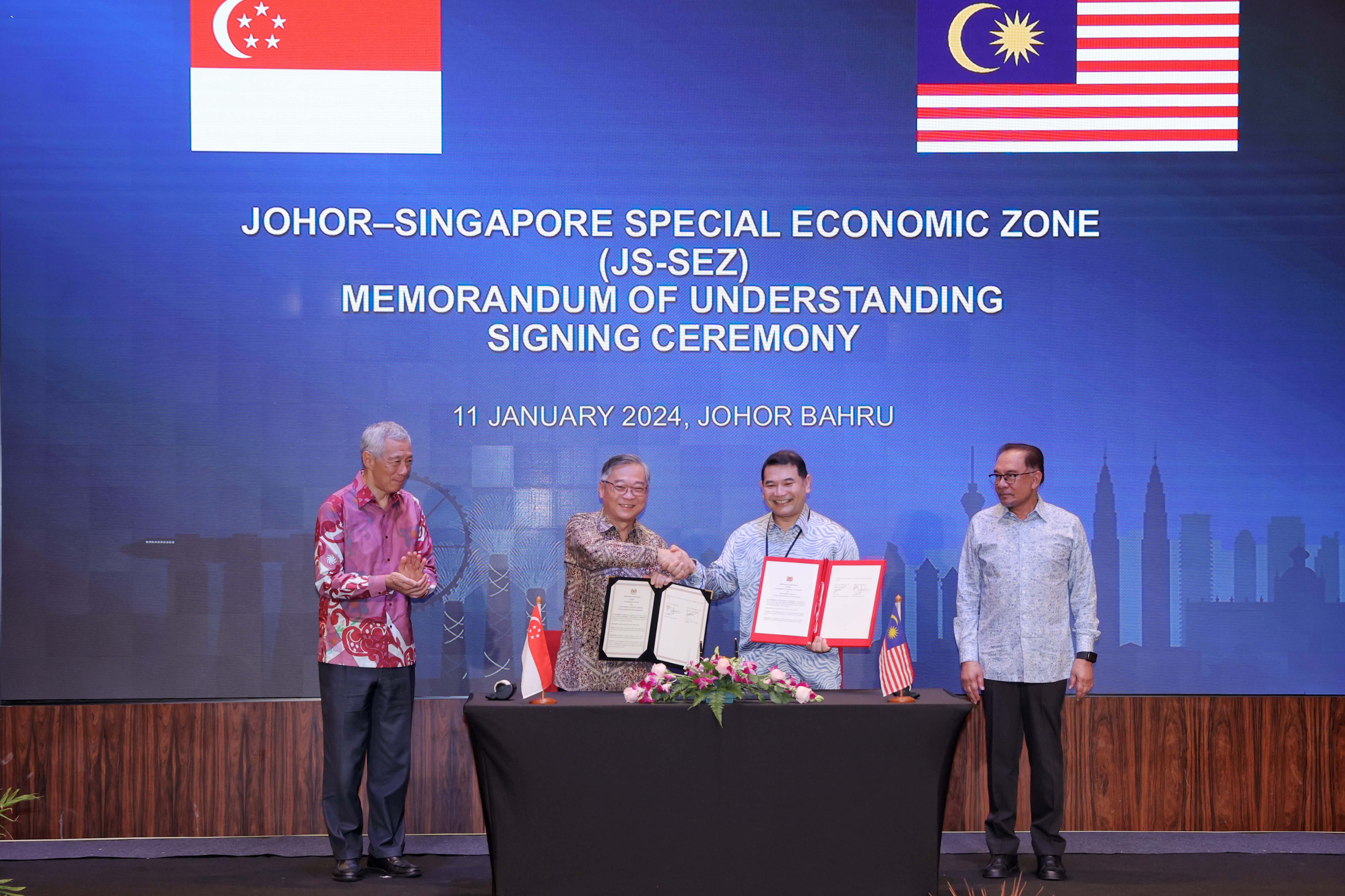 Four men in suits shake hands at JS-SEZ signing ceremony. Singapore & Malaysia flags displayed.