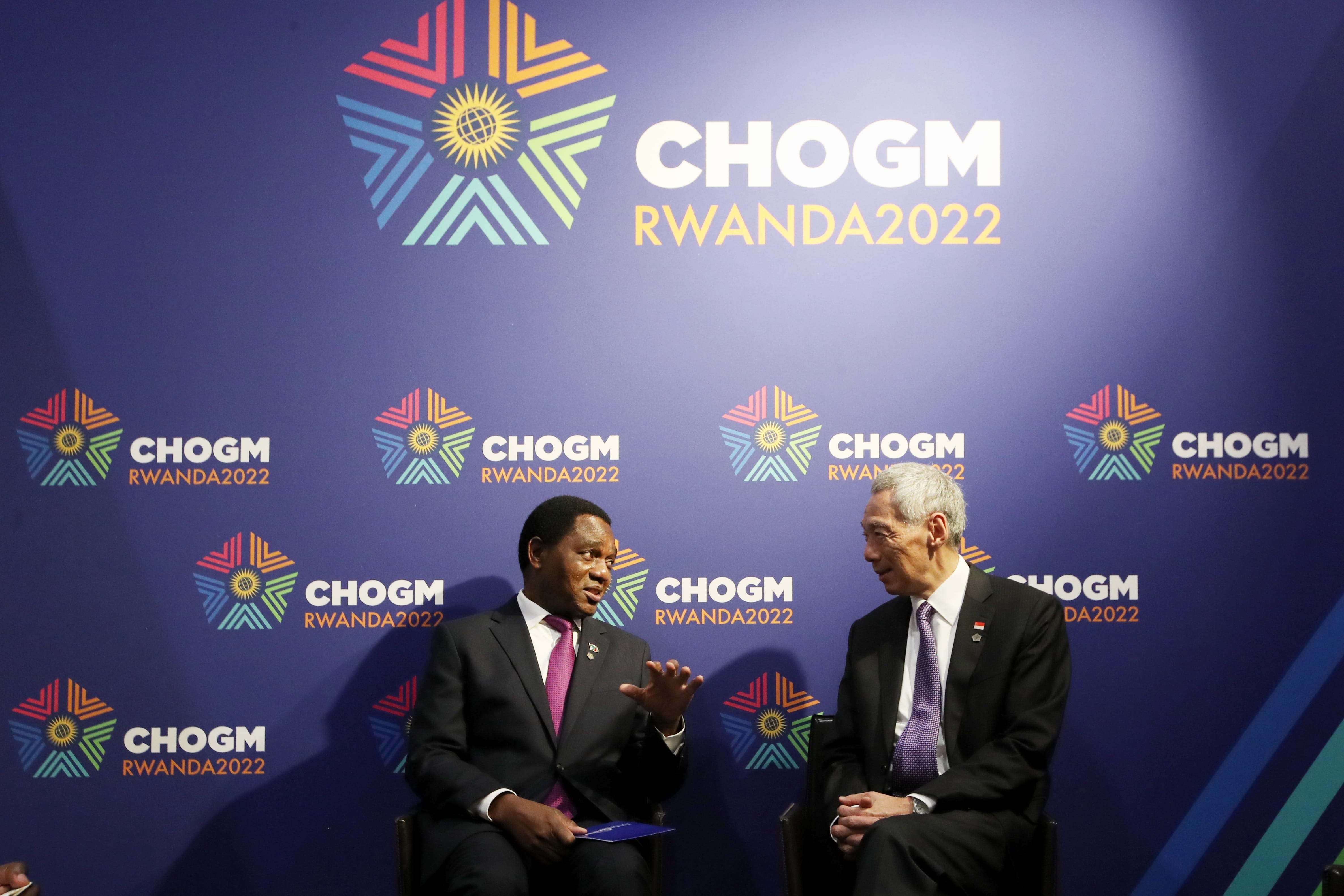 Two men in suits sit before a CHOGM RWANDA2022 backdrop. One gestures as if speaking.