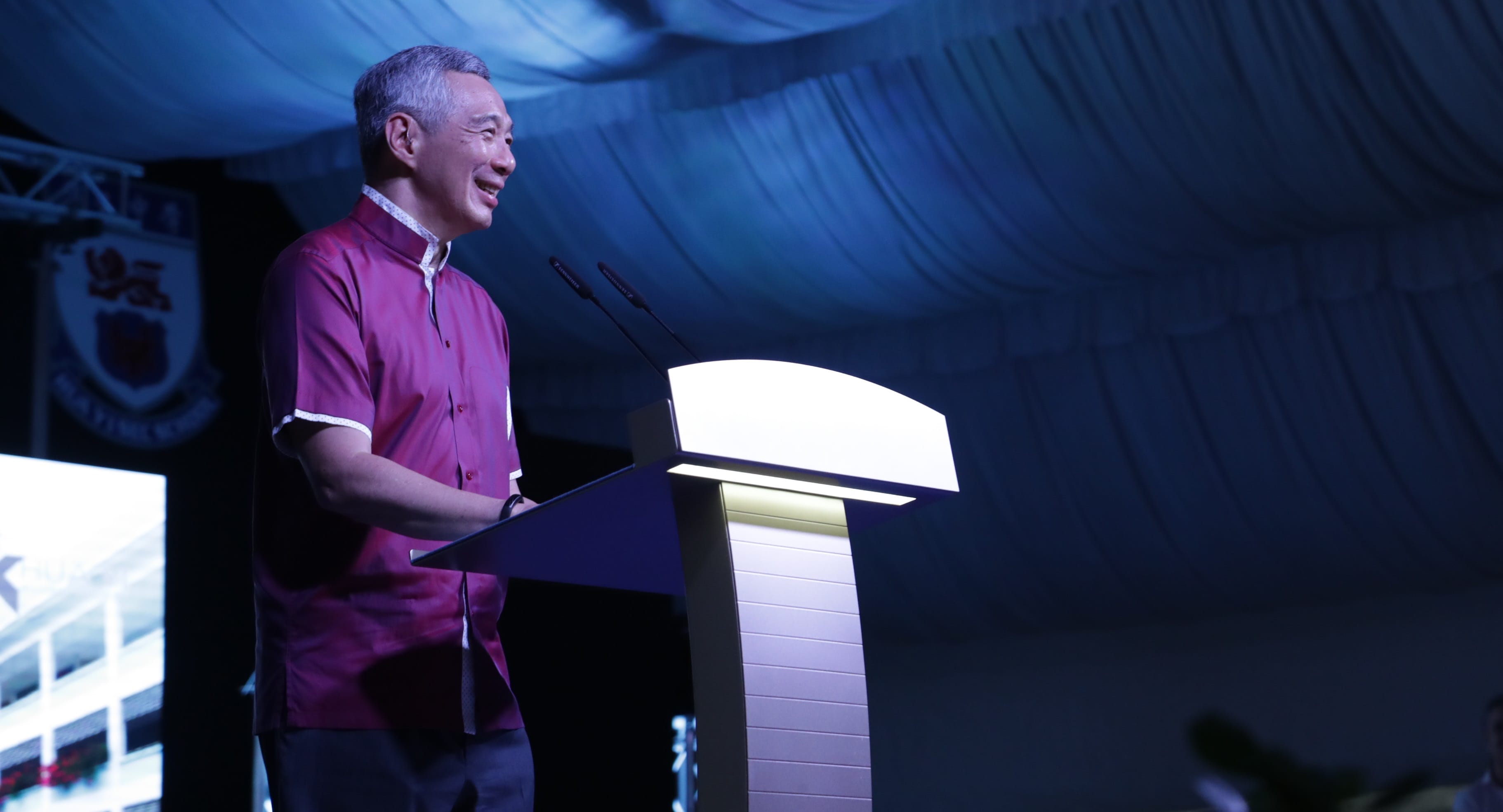 Lee Hsien Loong at a lit podium with Sennheiser mics, speaking under blue draped ceiling.