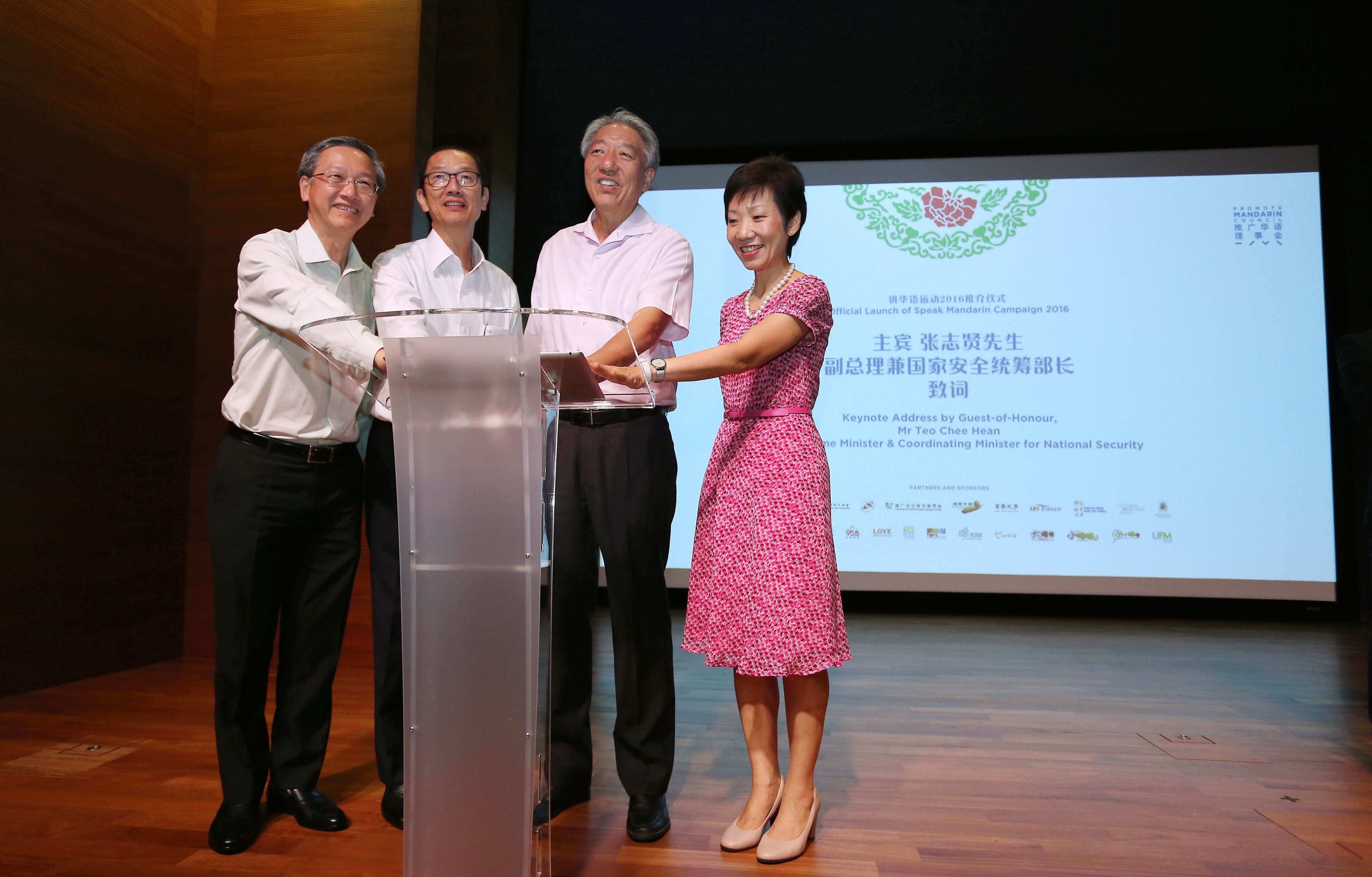 Four people place hands on lectern. Text projected behind reads "Speak Mandarin Campaign 2016".