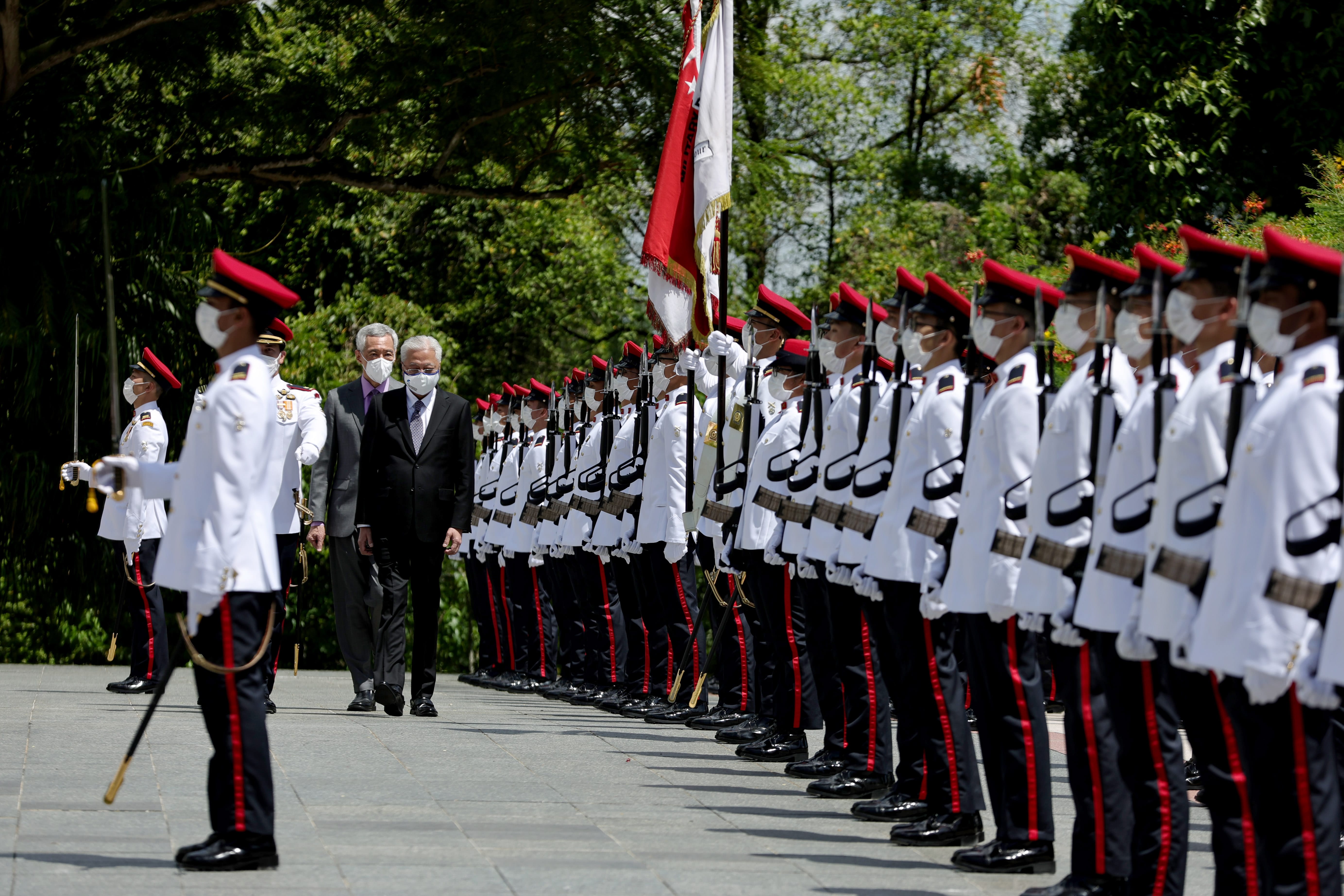 Two men in suits walk past honor guard in white uniforms with red hats and masks. Flags in background.