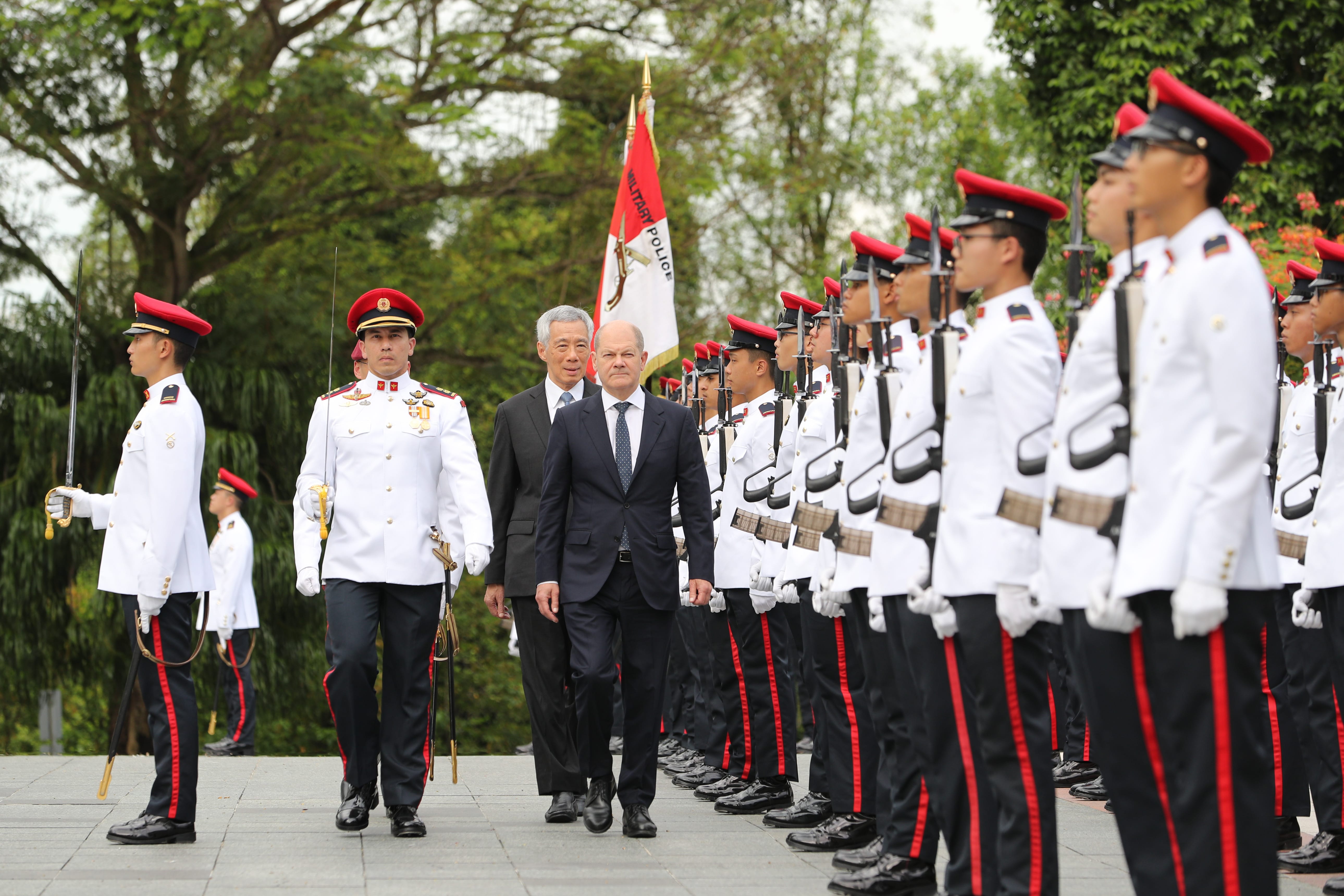 Olaf Scholz and Lee Hsien Loong walk alongside honor guard in white uniforms and red caps.