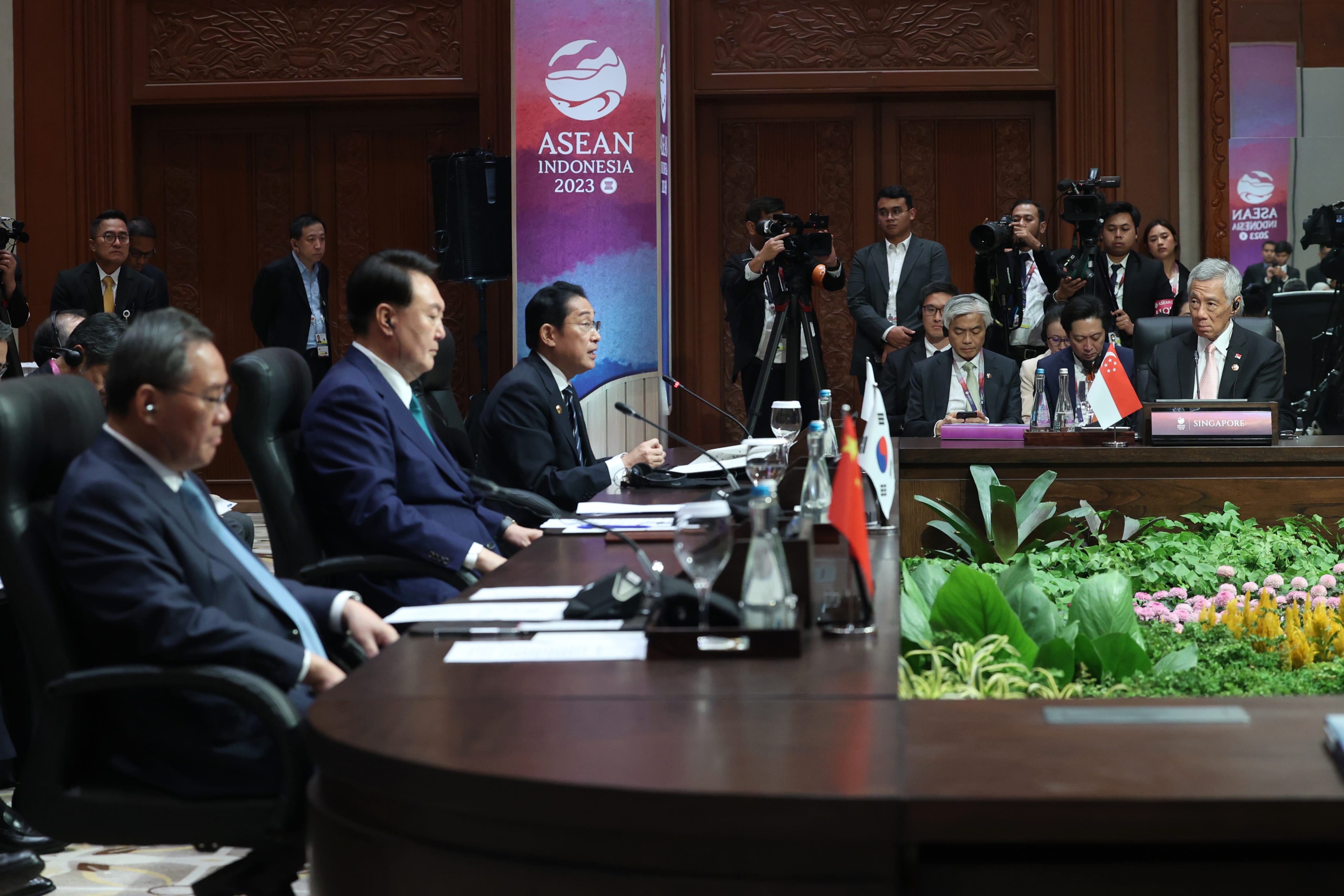 ASEAN Indonesia 2023 summit with world leaders at long conference table, flags of South Korea, Singapore. Cameras pointed.