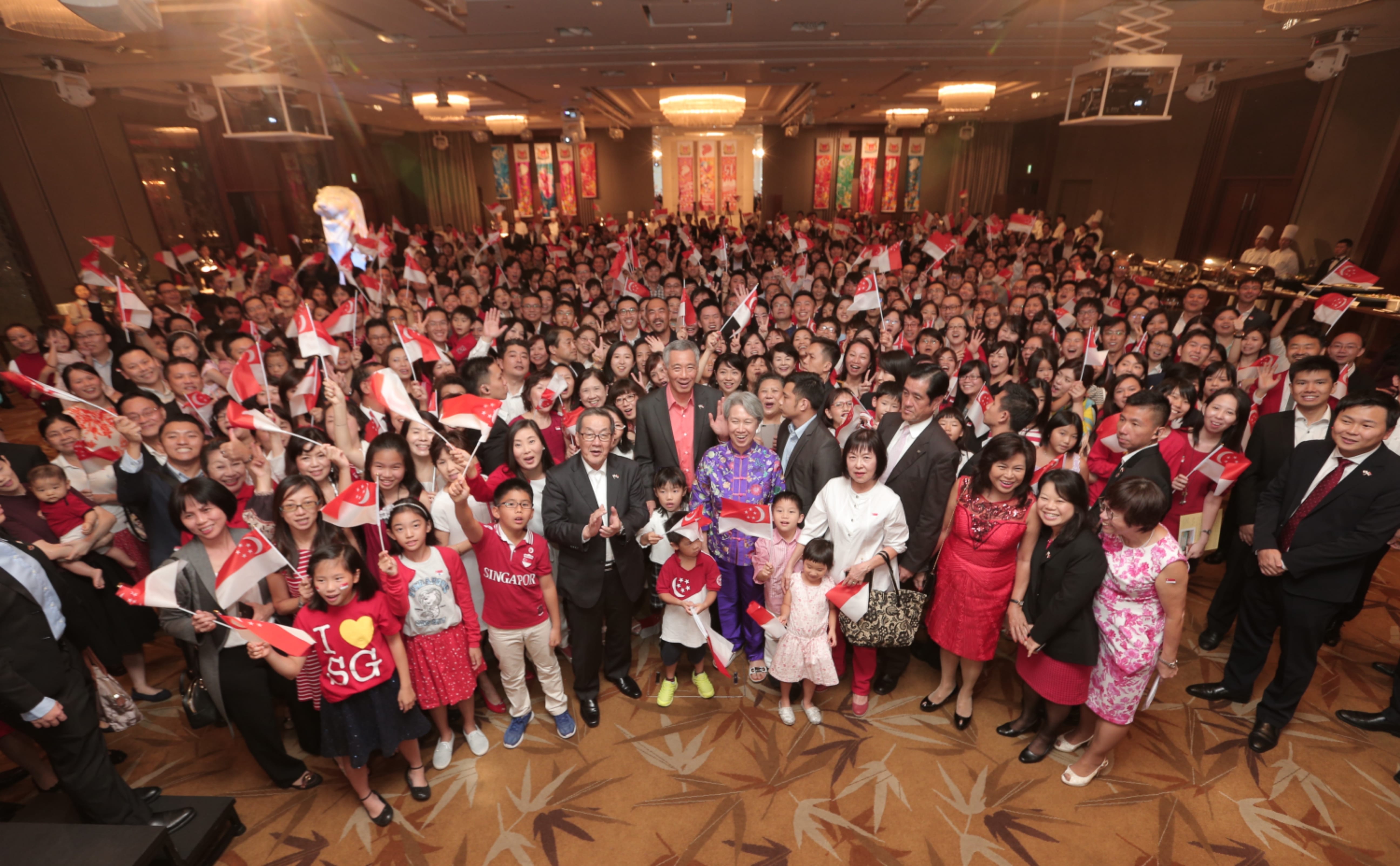 Large group holding Singapore flags in a banquet hall; Lee Hsien Loong visible.