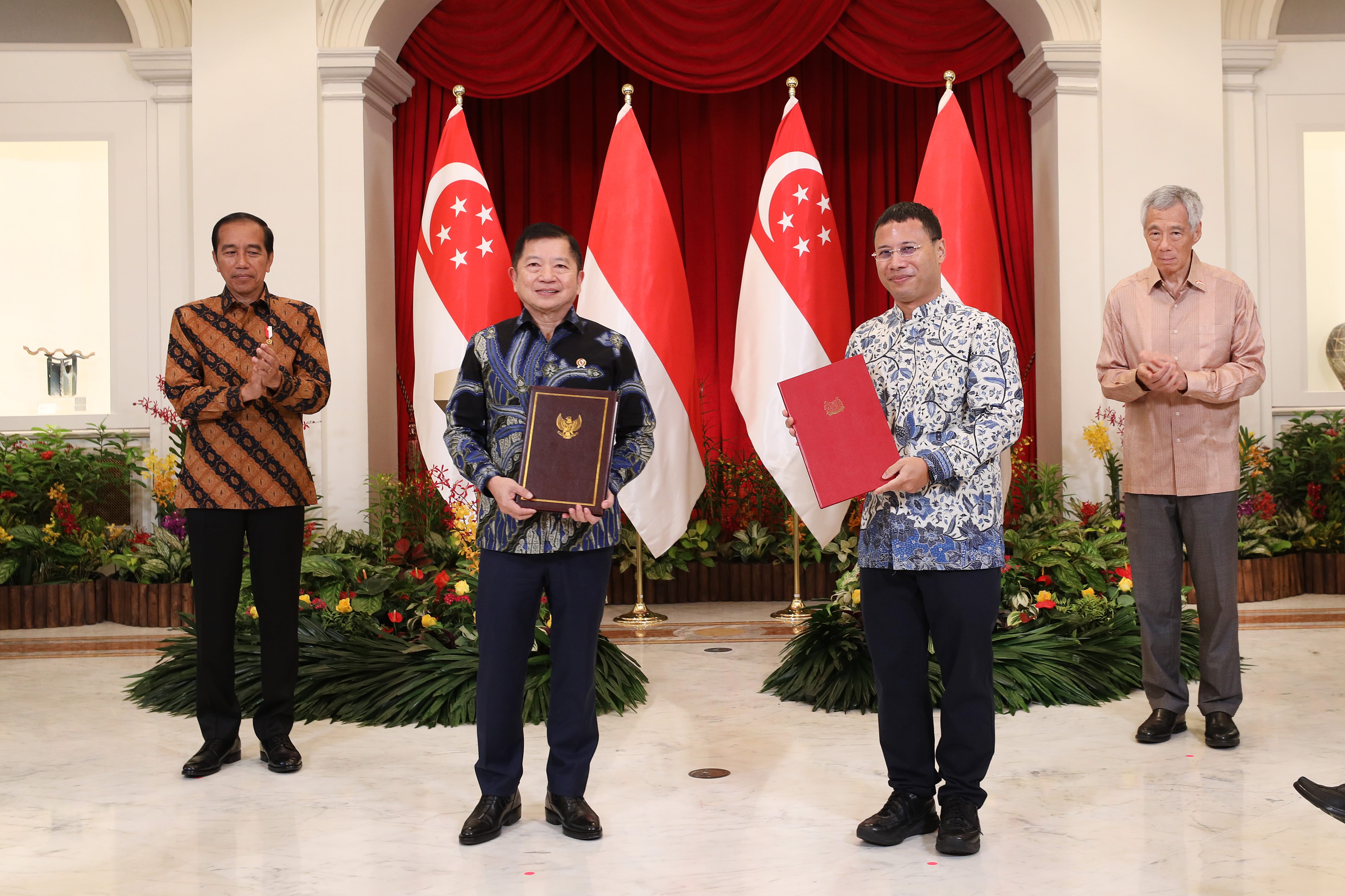 Four men in suits holding documents stand before flags. Two wear batik shirts.
