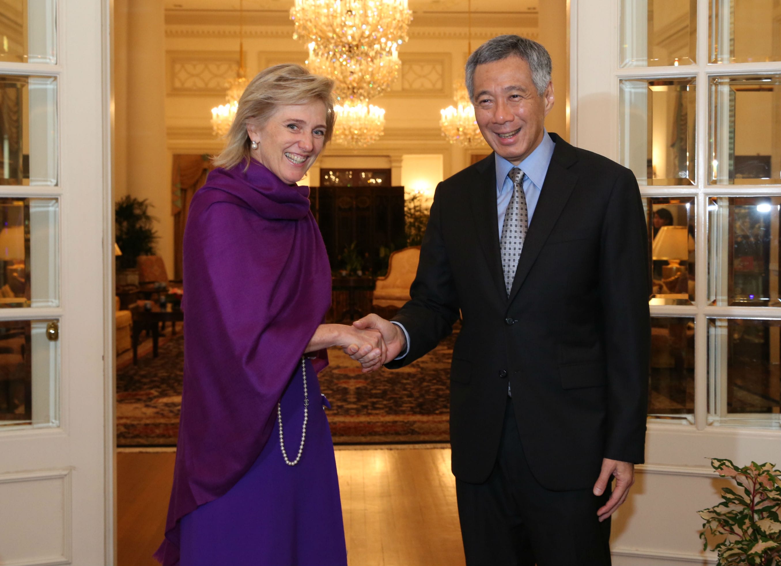Woman in purple shaking hands with Lee Hsien Loong in a suit, indoors with chandeliers.