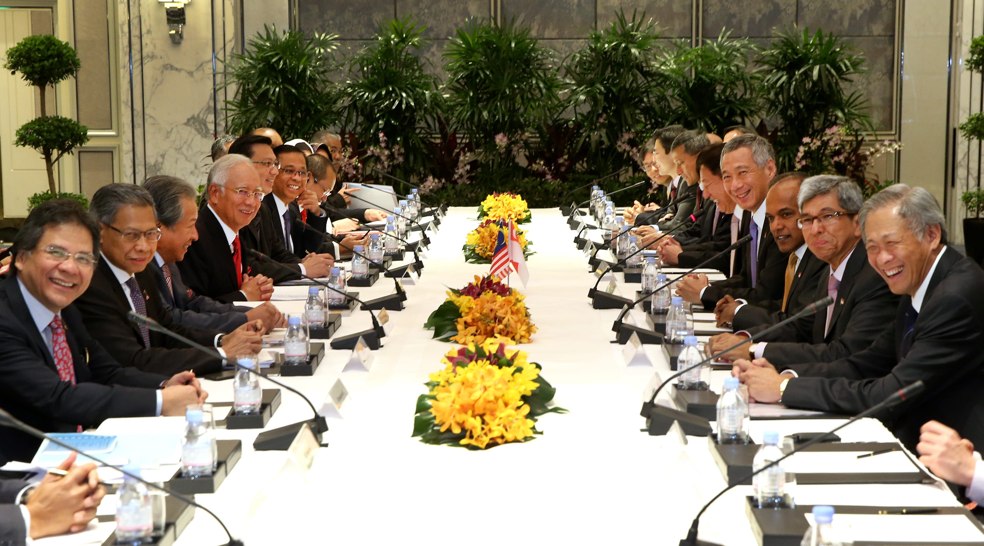 People sit at a long table with microphones, flags, water bottles, and flowers.
