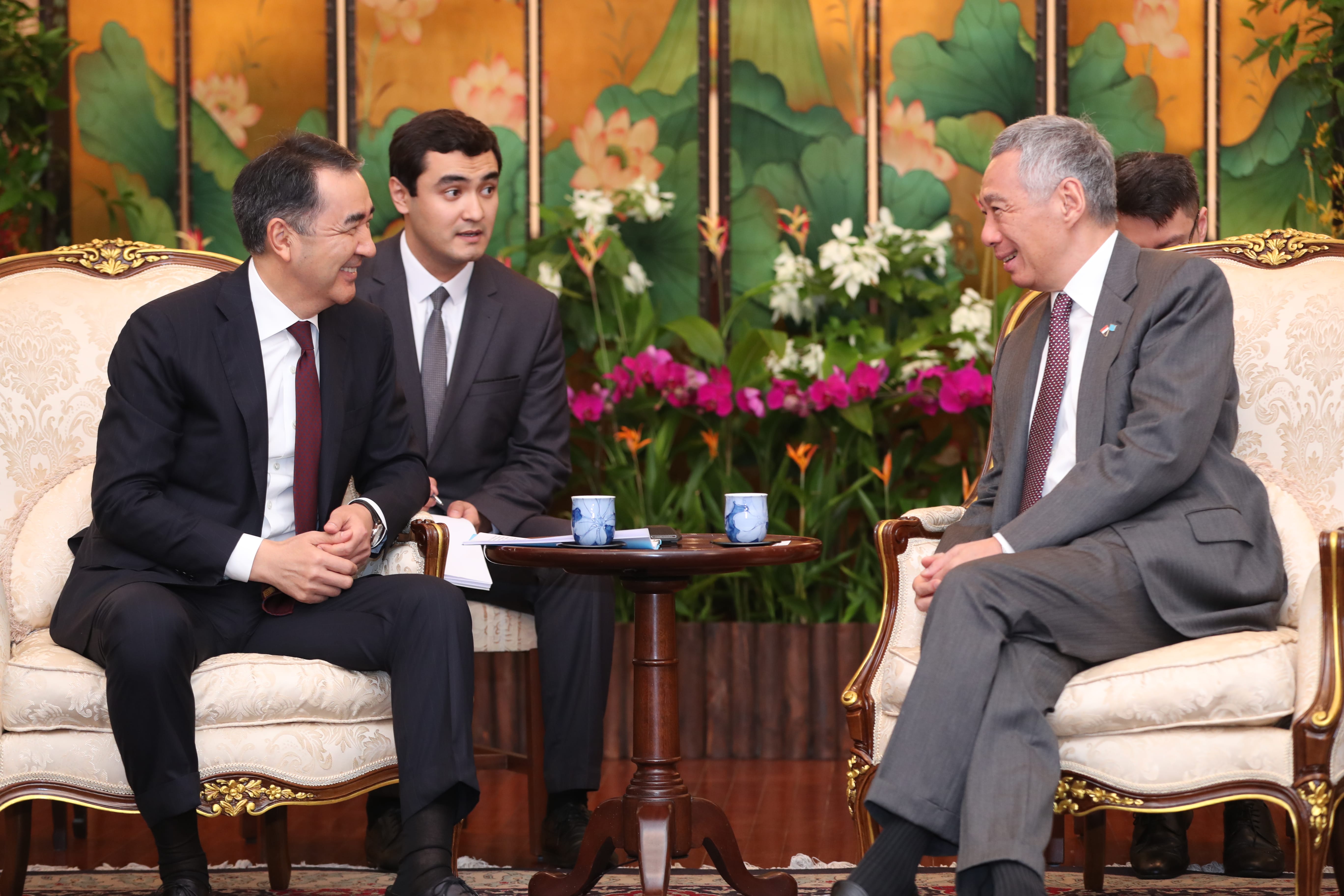 Three men in suits sit in ornate chairs, talking. Flowers decorate background.