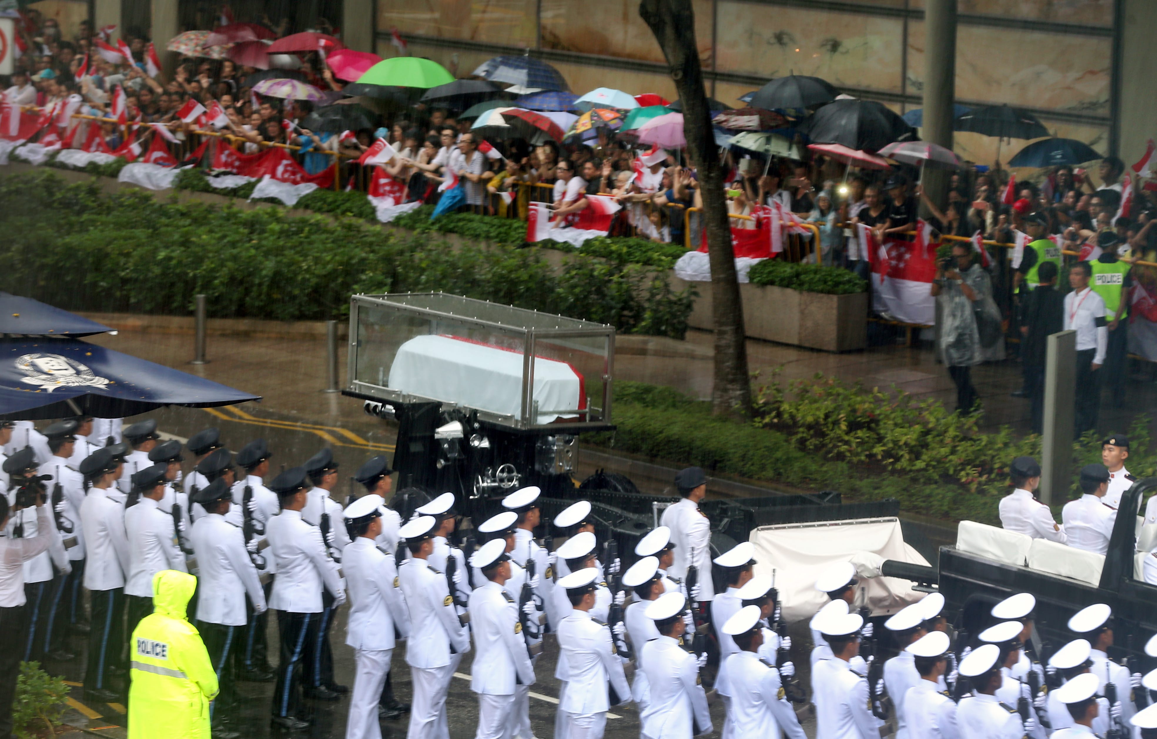 Rainy street scene with casket on vehicle, honor guard, & crowds holding Singapore flags.