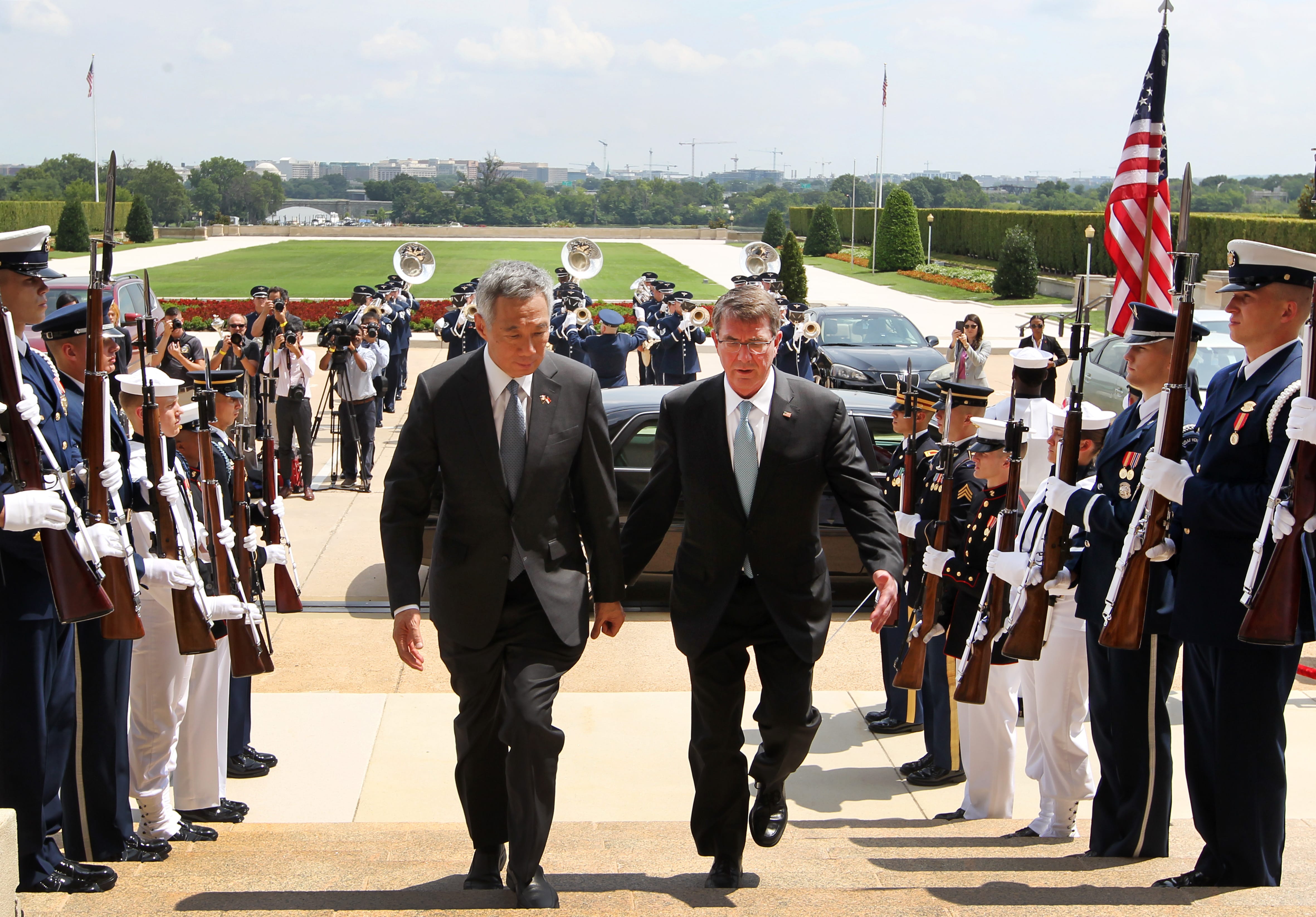 Two men in suits descend steps between honor guard holding rifles; a band plays in the background.