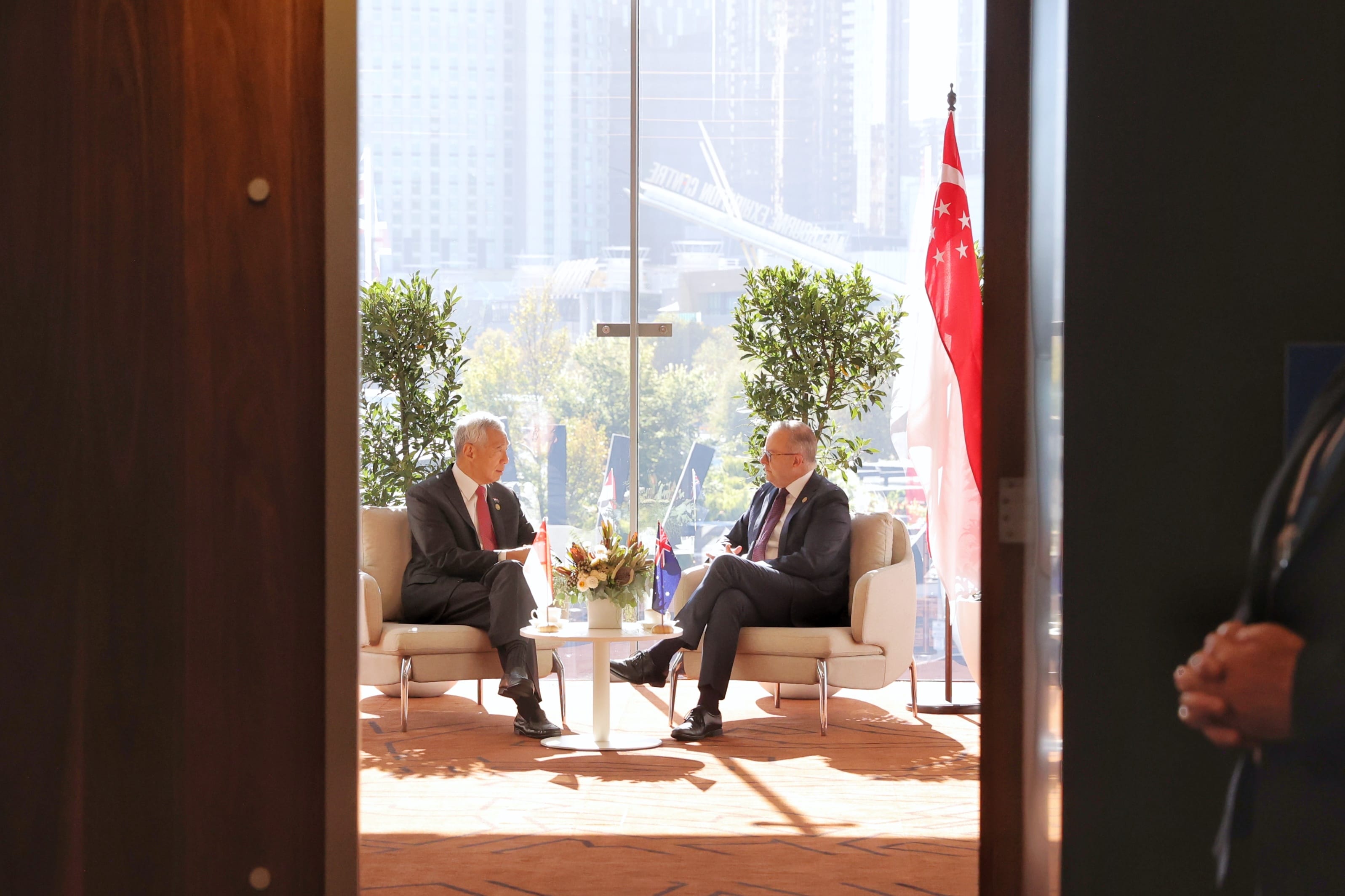 Two men in suits seated indoors, Singapore flag, city skyline in the background.