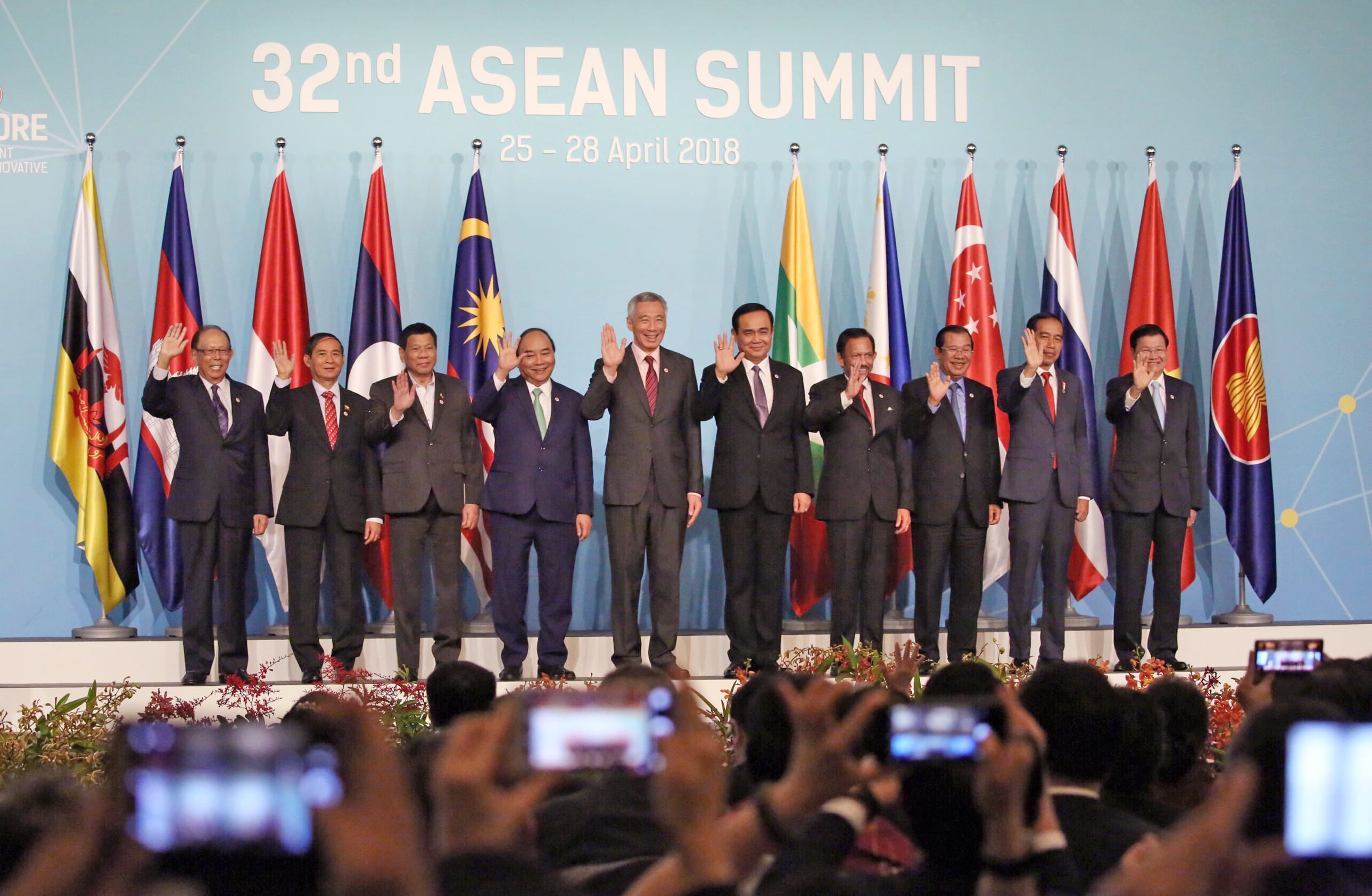 Group of people in suits wave in front of national flags at "32nd ASEAN SUMMIT." People in foreground taking photos.