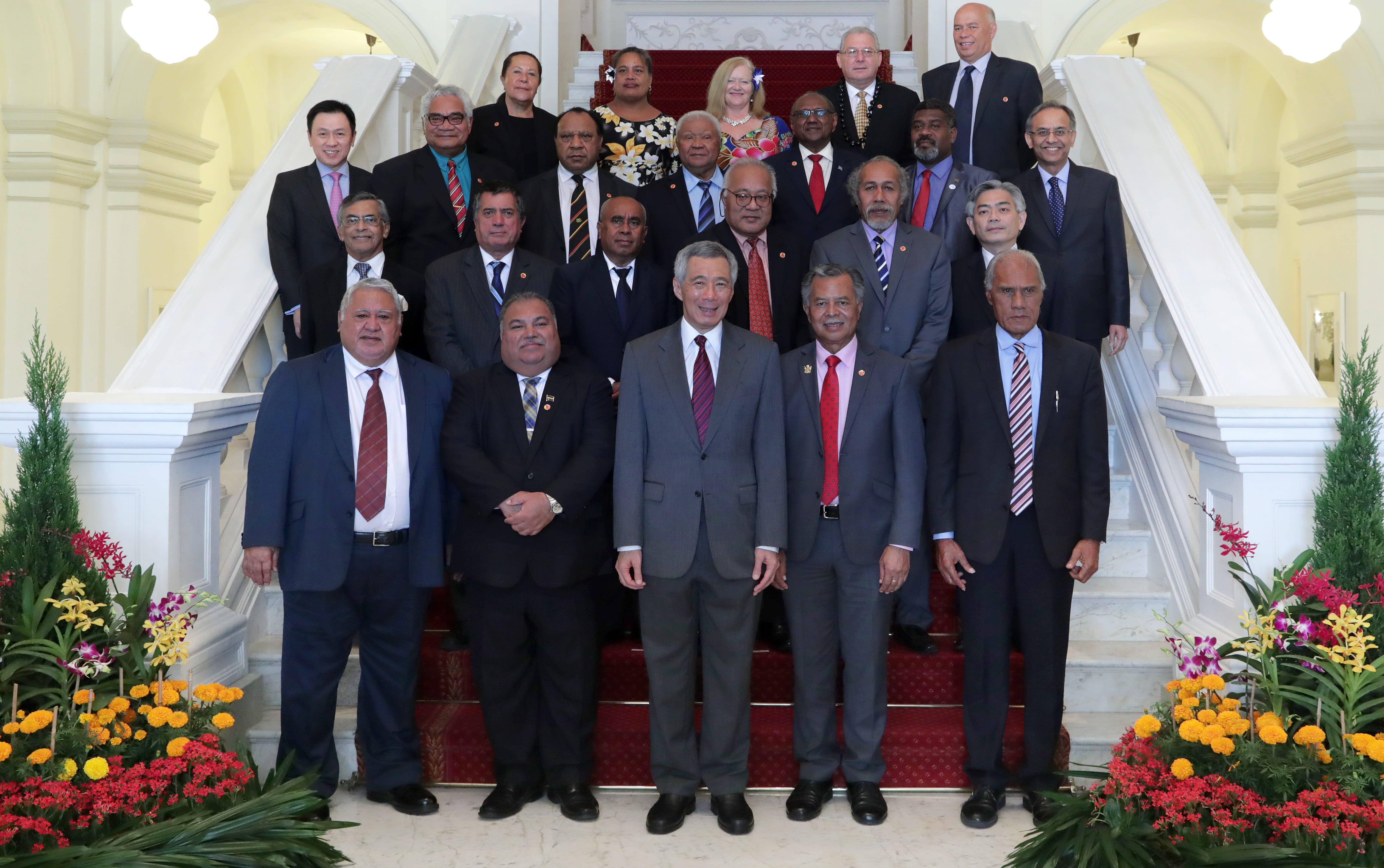 Group of diverse people in suits posing on staircase with red carpet and flower arrangements.