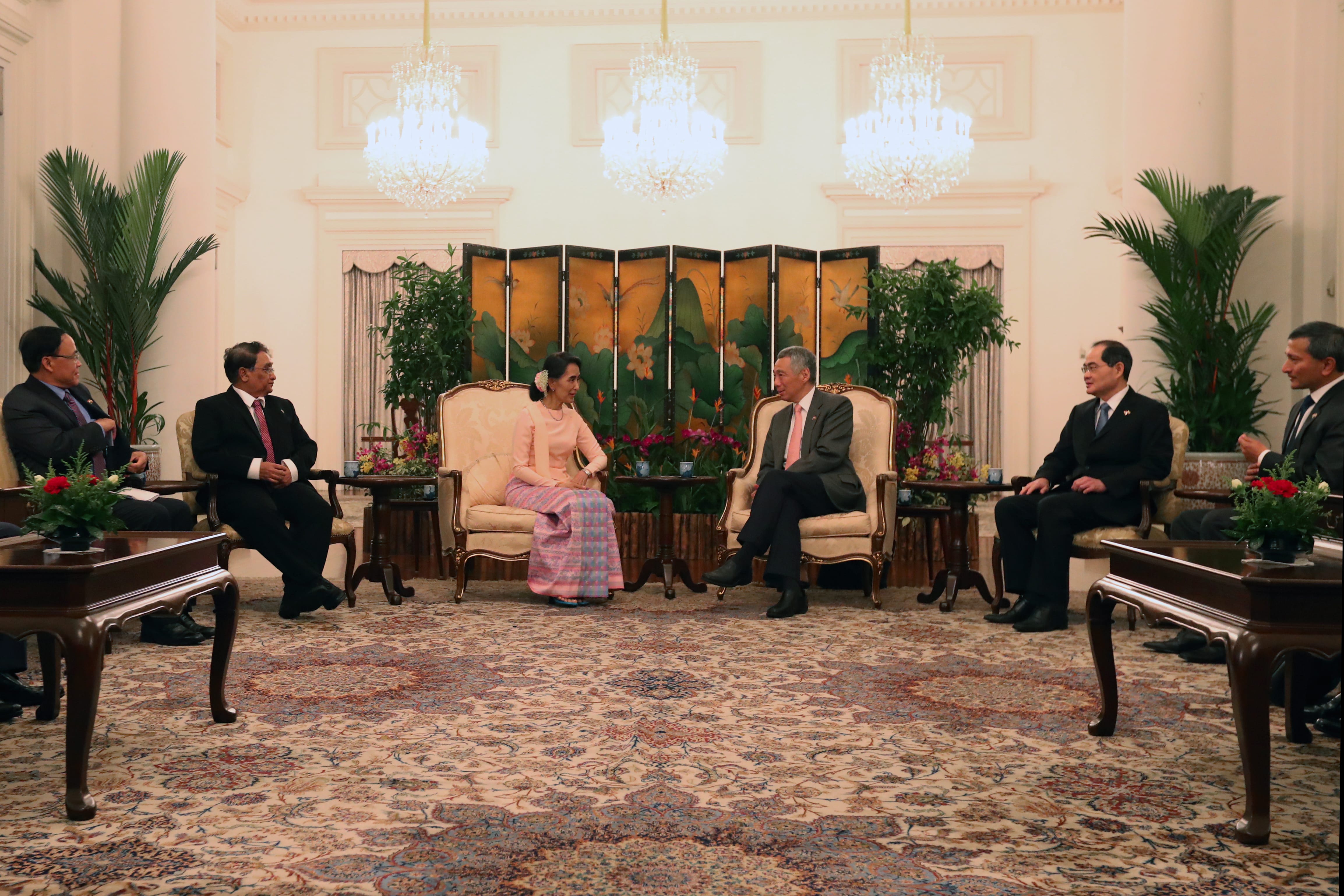 Aung San Suu Kyi and Lee Hsien Loong seated, surrounded by others in suits, in an ornate room.