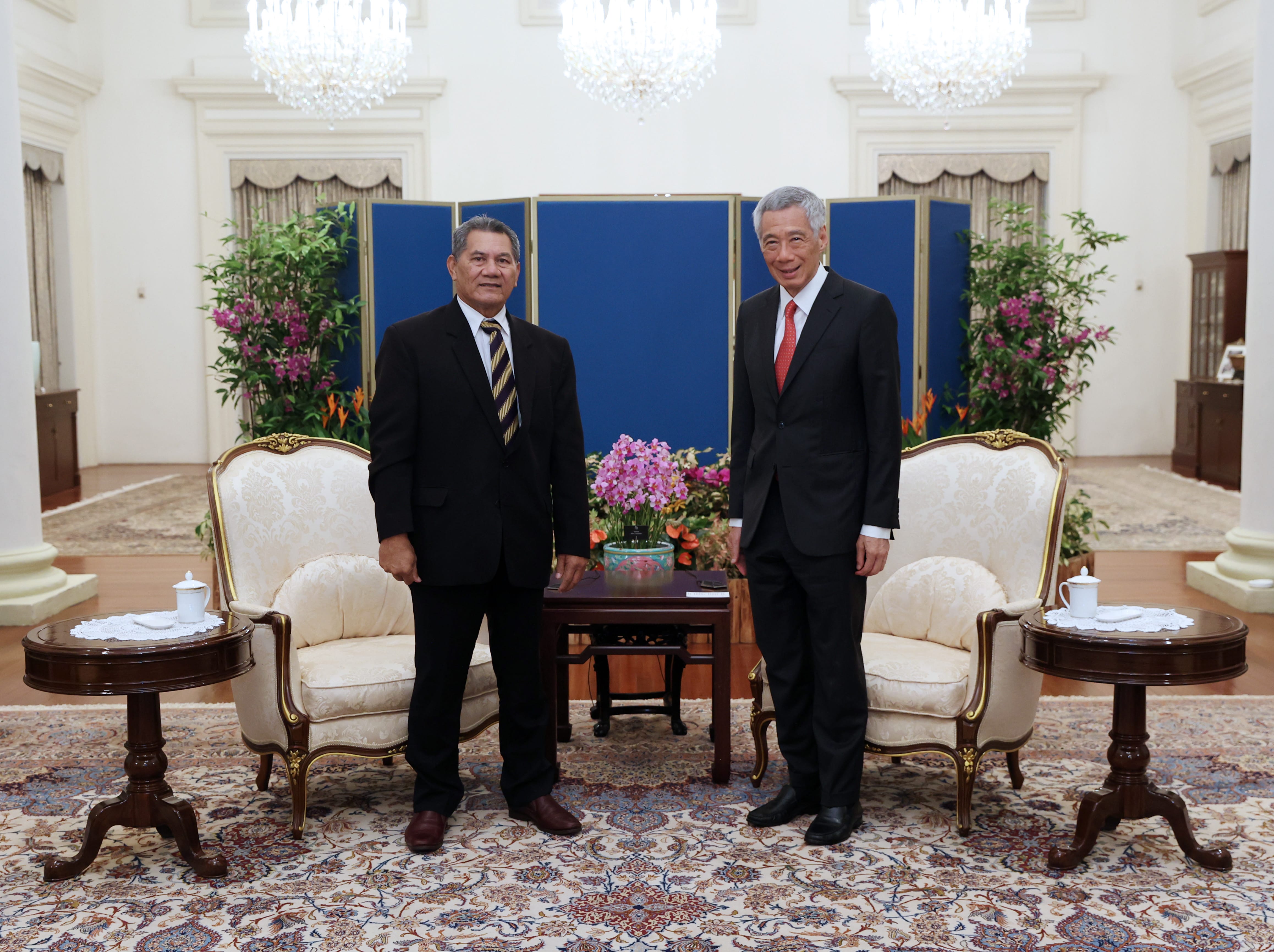 Two men in suits pose in a room with ornate chairs, tables, and floral arrangements.