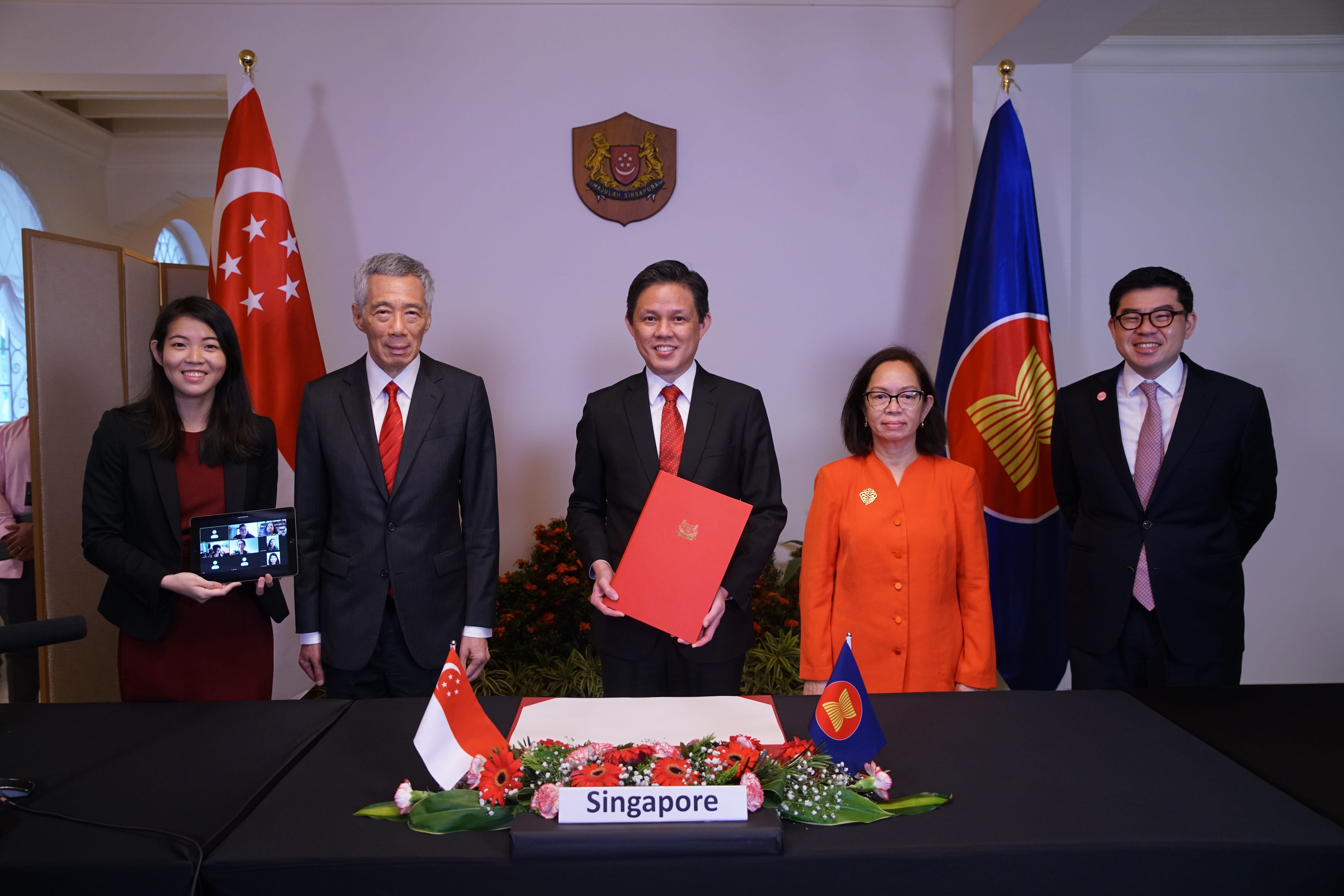 Five people and a tablet stand before Singapore and ASEAN flags and seal at a signing event.