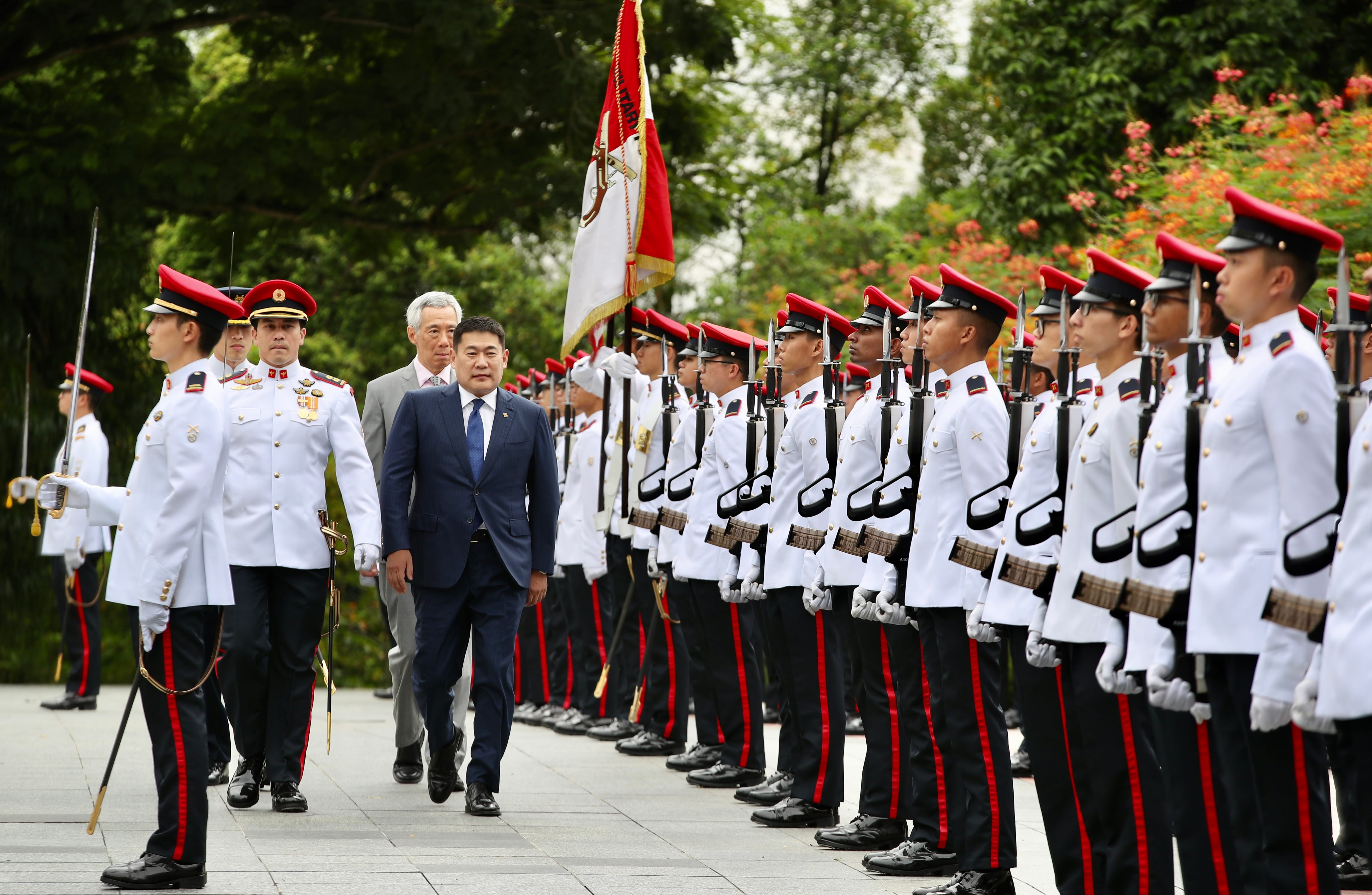 Line of soldiers in red-trimmed white uniforms face forward, dignitaries walk alongside.