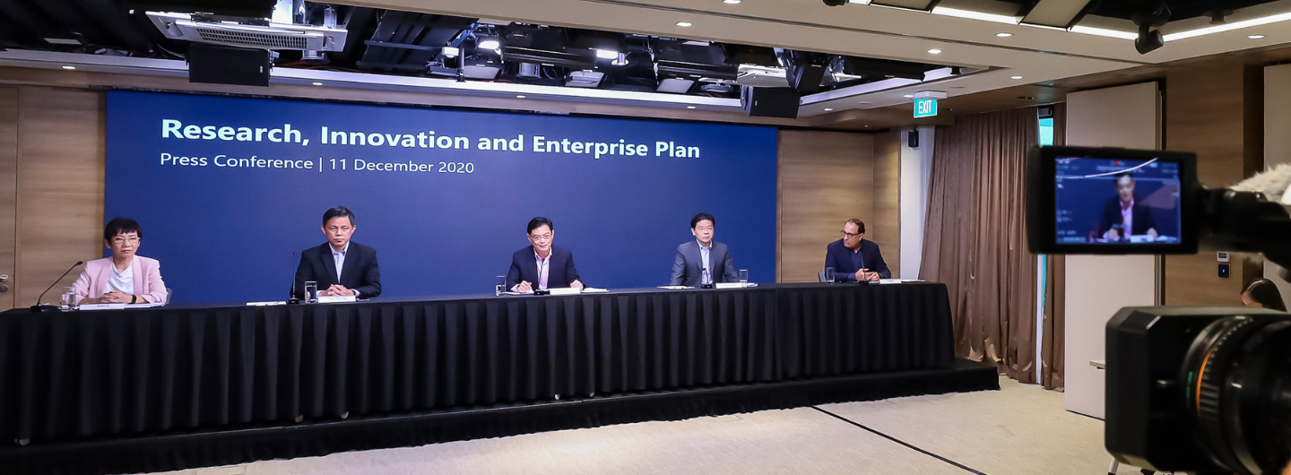 Five people sit behind a table at a "Research, Innovation and Enterprise Plan" press conference; a camera is recording.