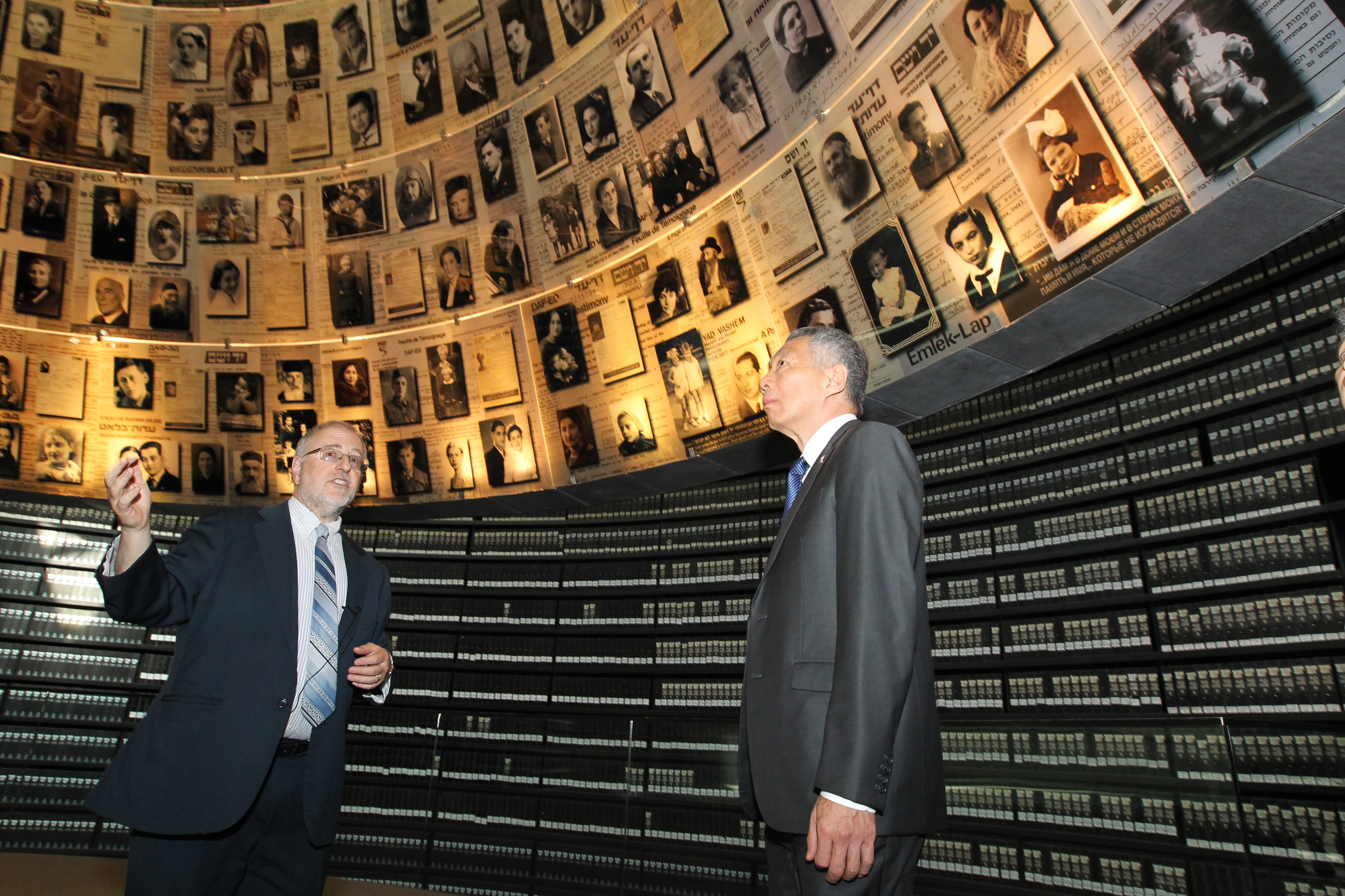 Two men in suits stand before a wall covered with photos and documents above rows of books.