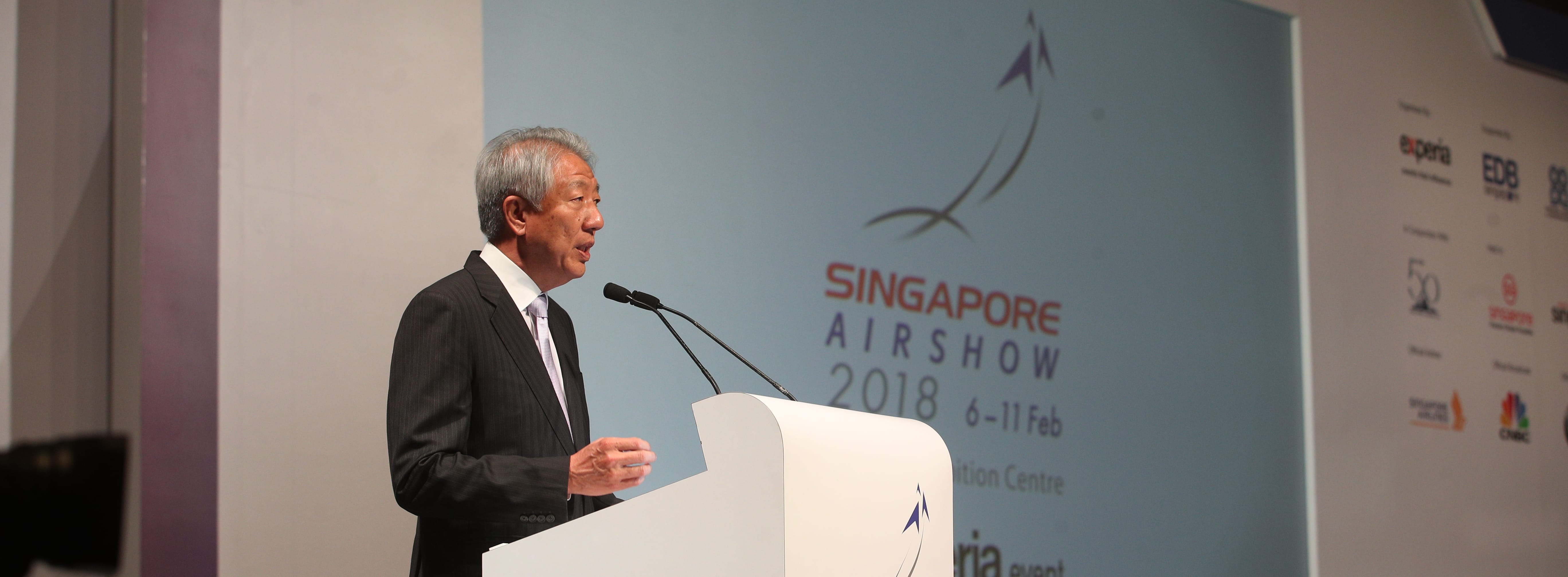 Man at podium with microphones, Singapore Airshow 2018 logo behind him, sponsor logos visible to the right.