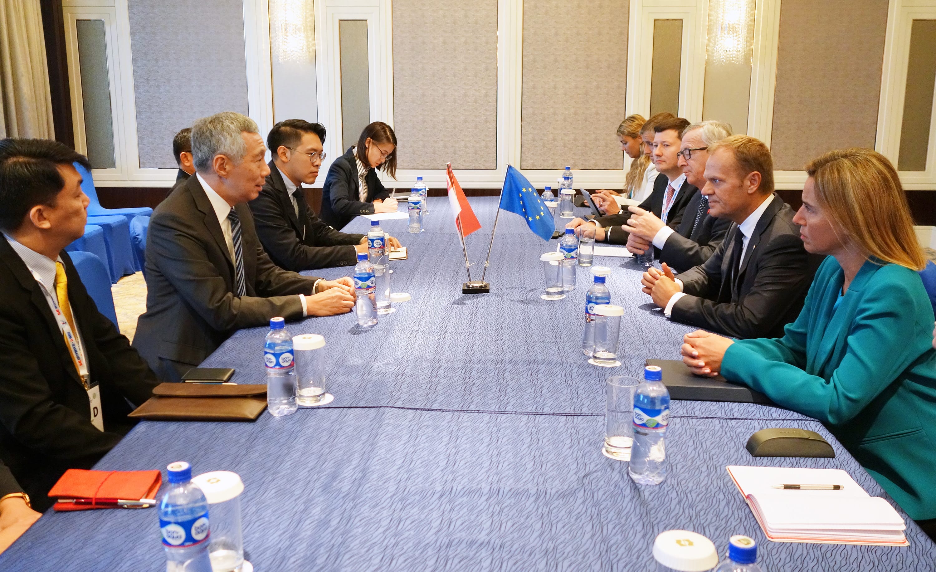 People sit at a conference table with Singapore and EU flags. Bottled water is on the table.