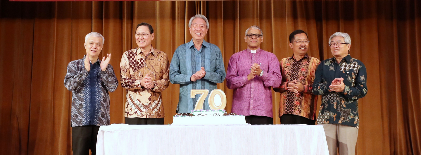 Six people applaud behind a cake with "70" on a stage with brown curtains.