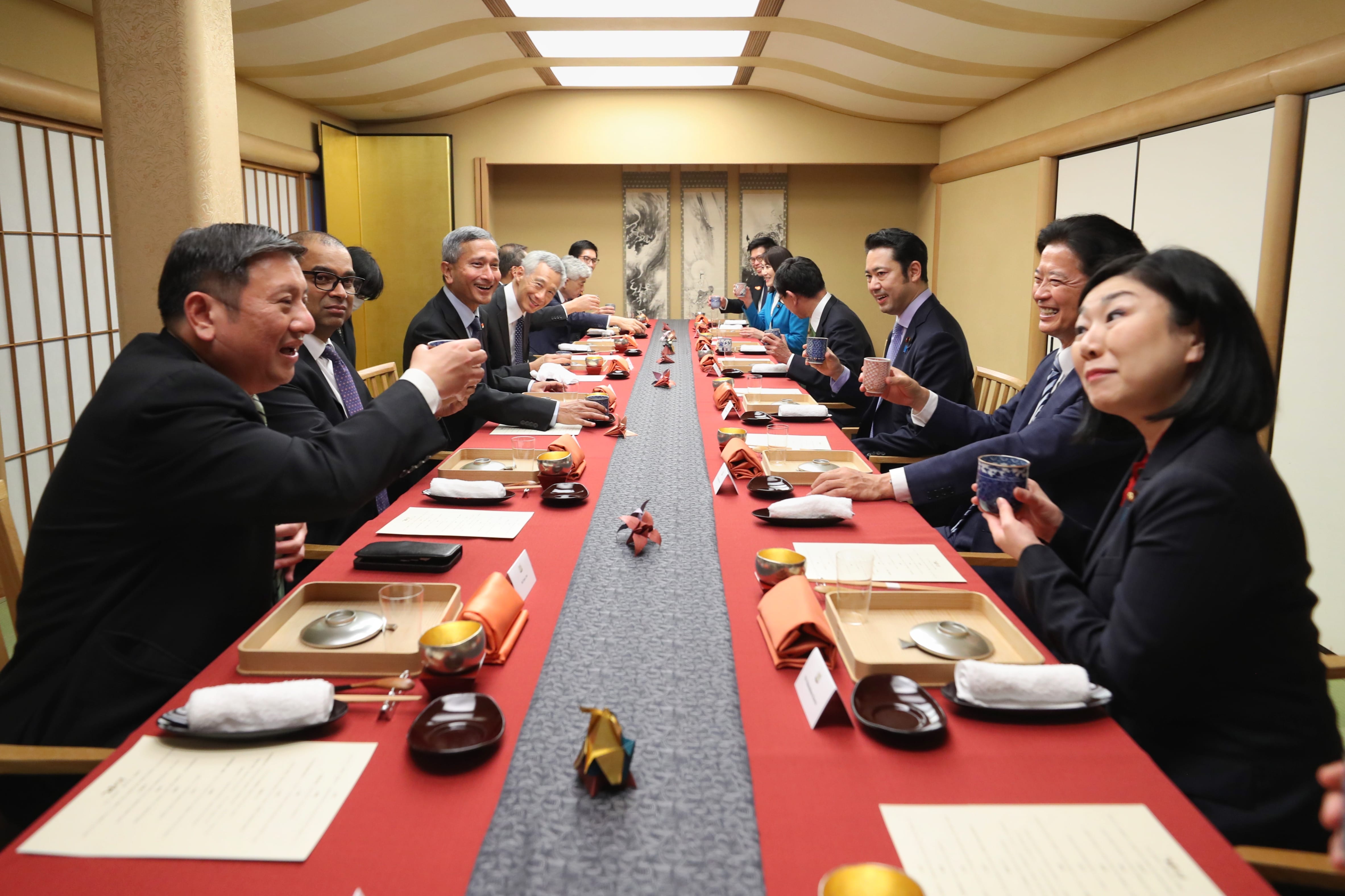 People at long table with red runner, toasting with cups in a bright room.