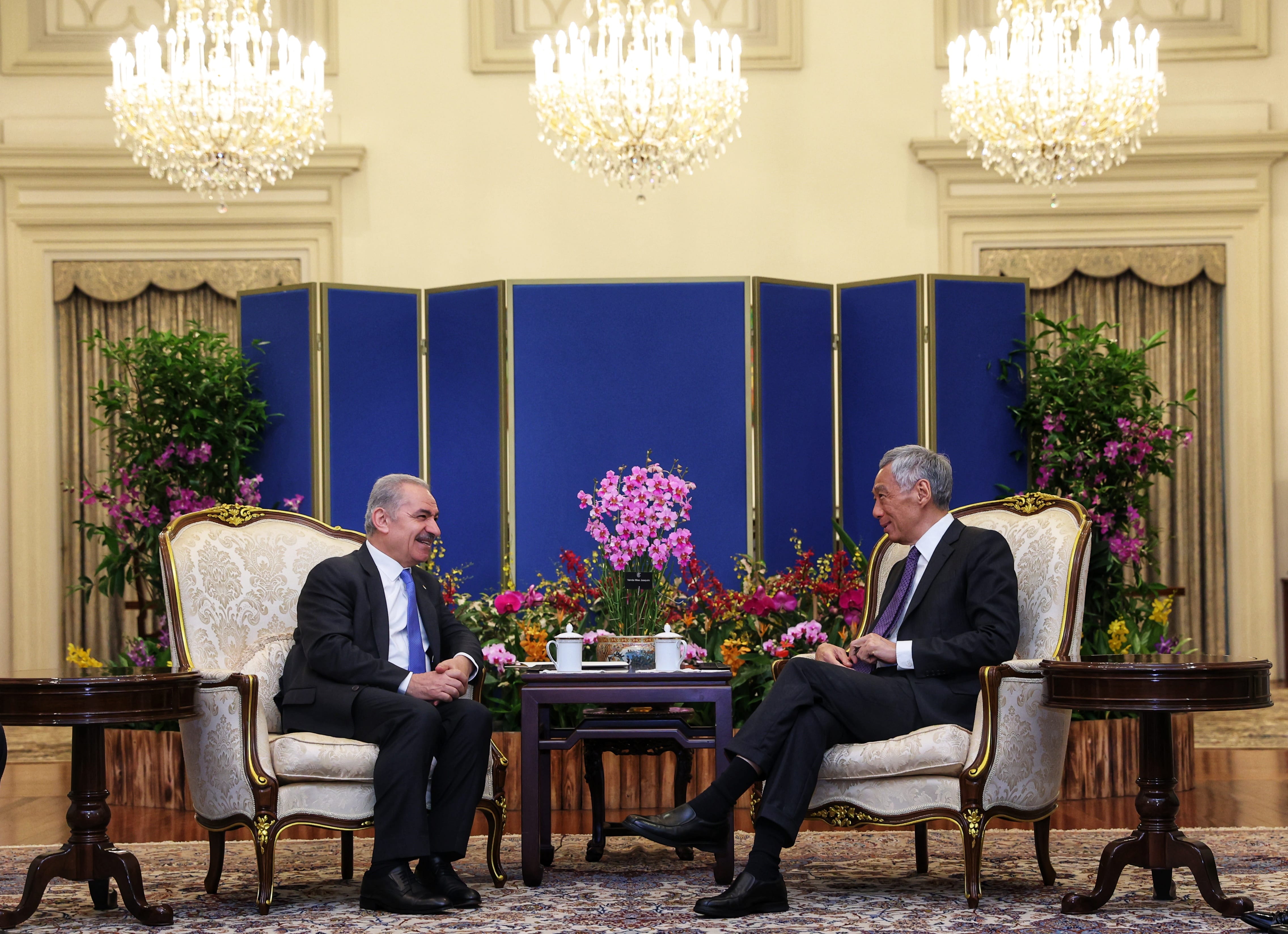 Two men seated in ornate chairs facing each other, flowers between them, chandeliers above.