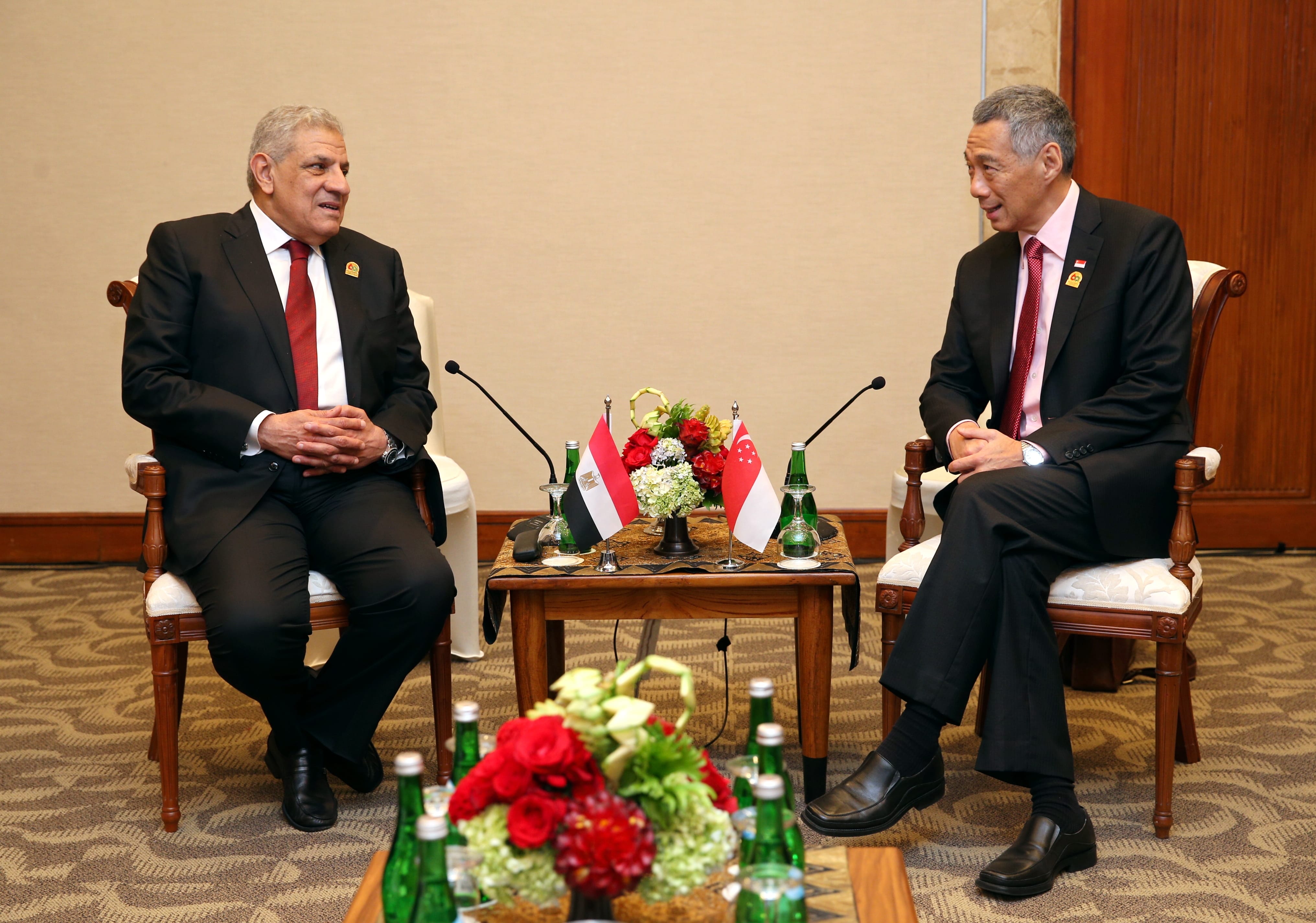 Two men in suits seated facing each other with flags of Egypt and Singapore on table.