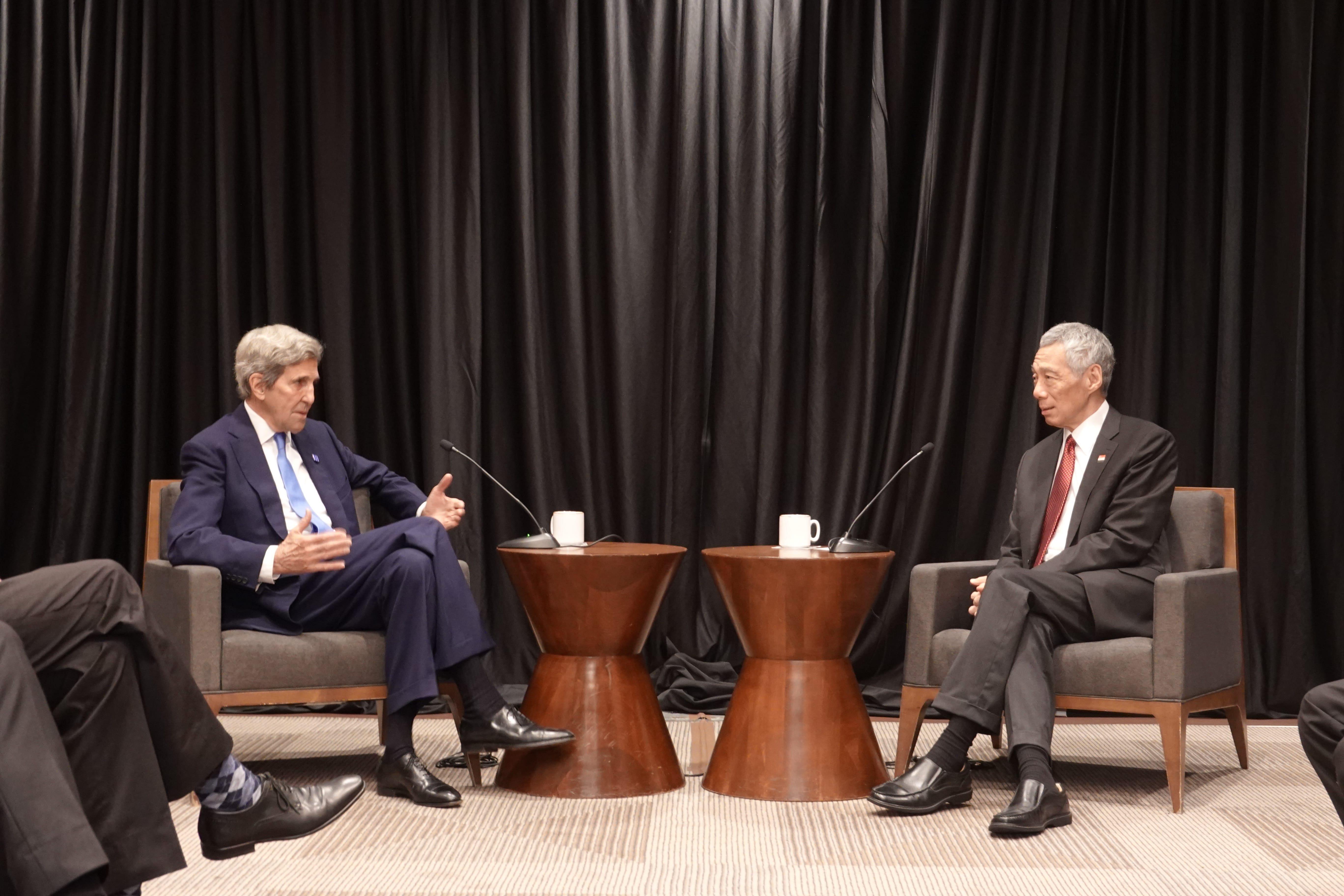 John Kerry and Lee Hsien Loong seated, speaking at a forum, mugs on hourglass tables.