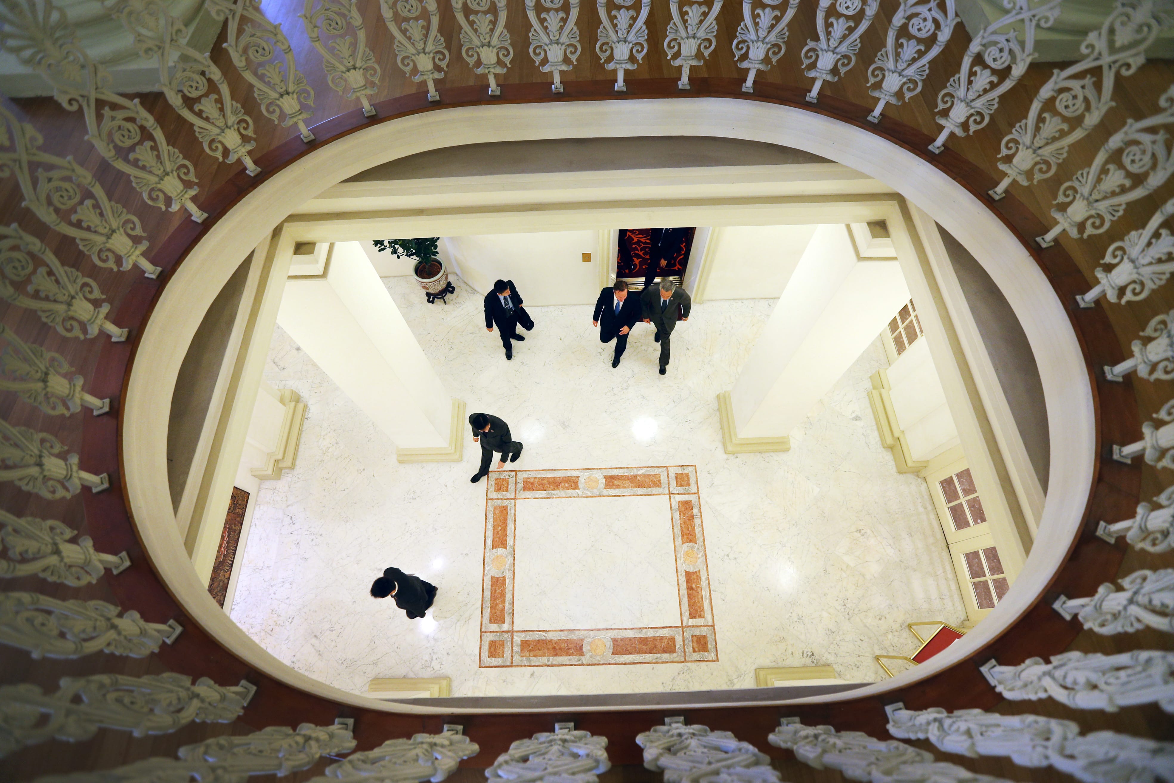 Looking down on people in suits walking across a marble floor, framed by ornate railing.
