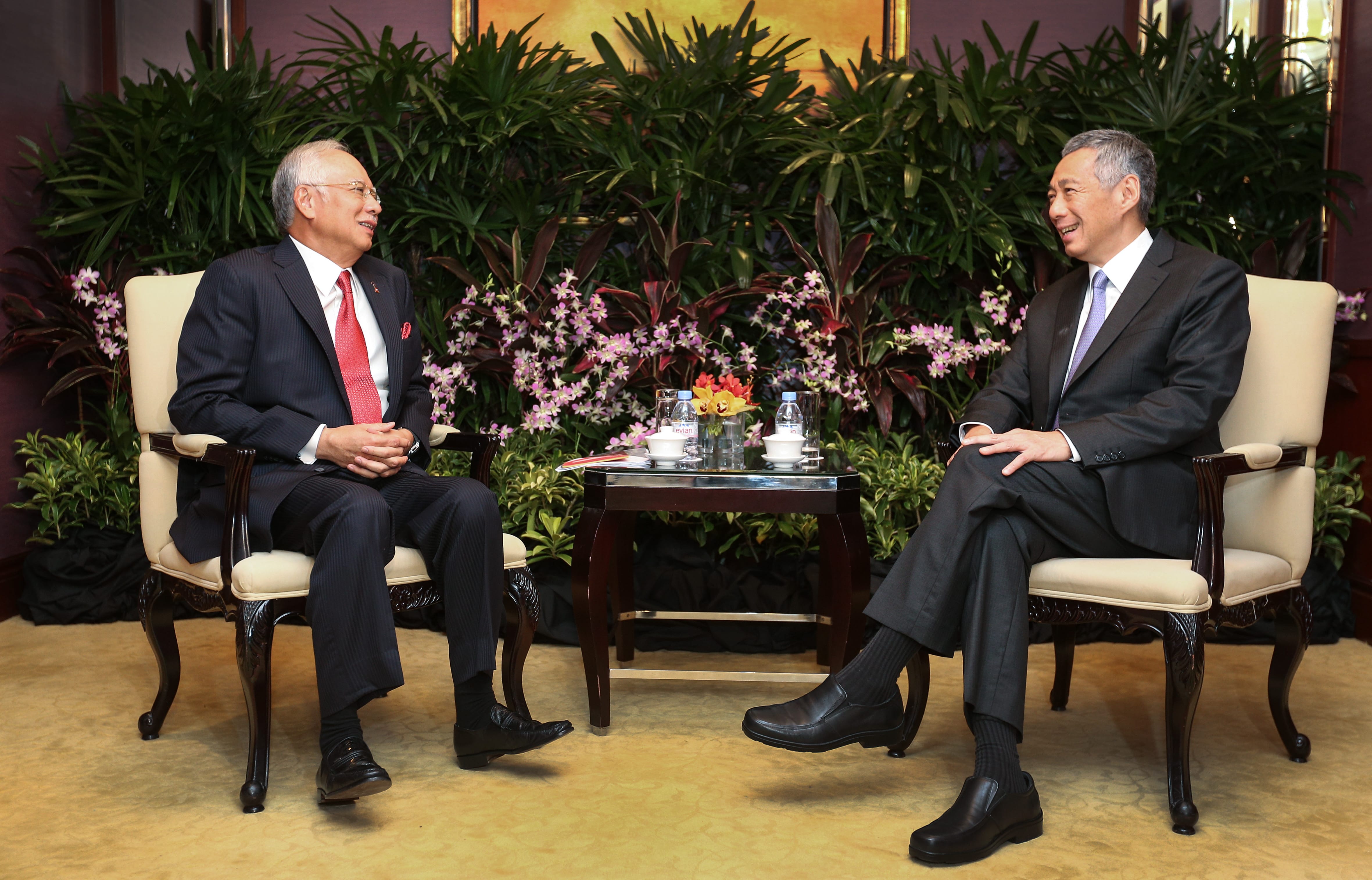 Two men in suits seated, facing each other, with plants and flowers in background.