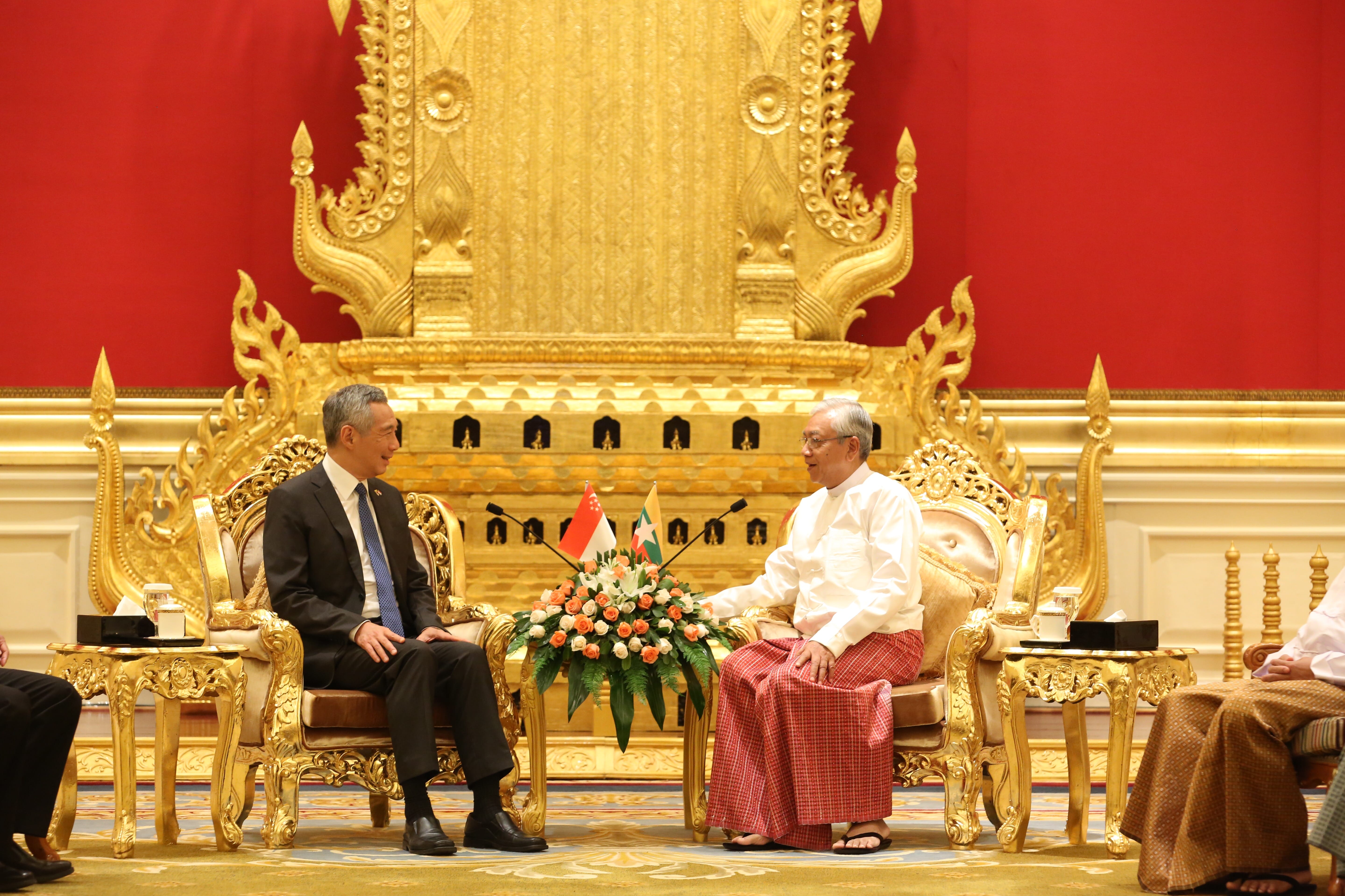 Two men sit in ornate gold chairs facing each other, flags and flowers between them.