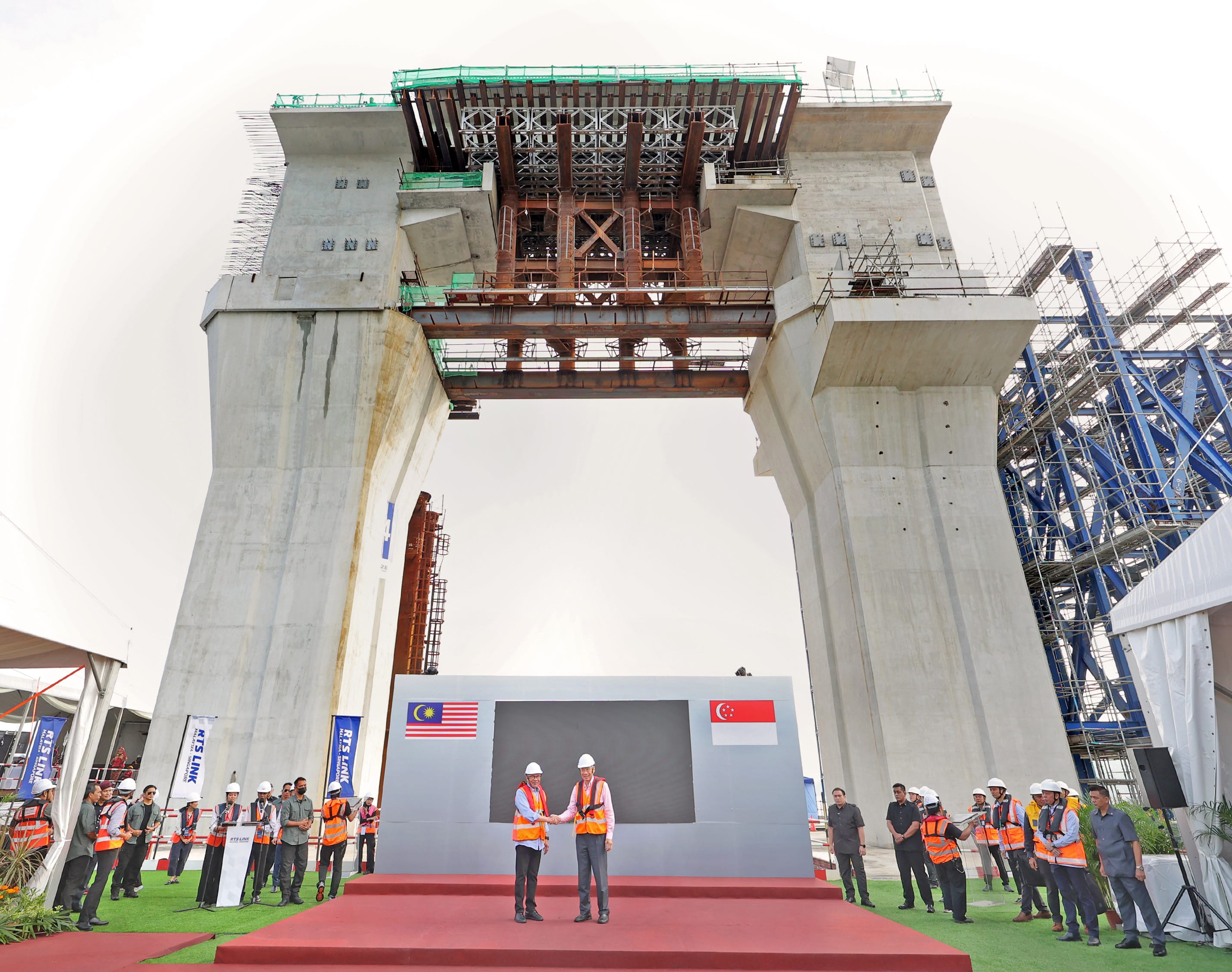 RTS Link construction: Two men shake hands on stage, Malaysian and Singaporean flags behind. Crowd present.