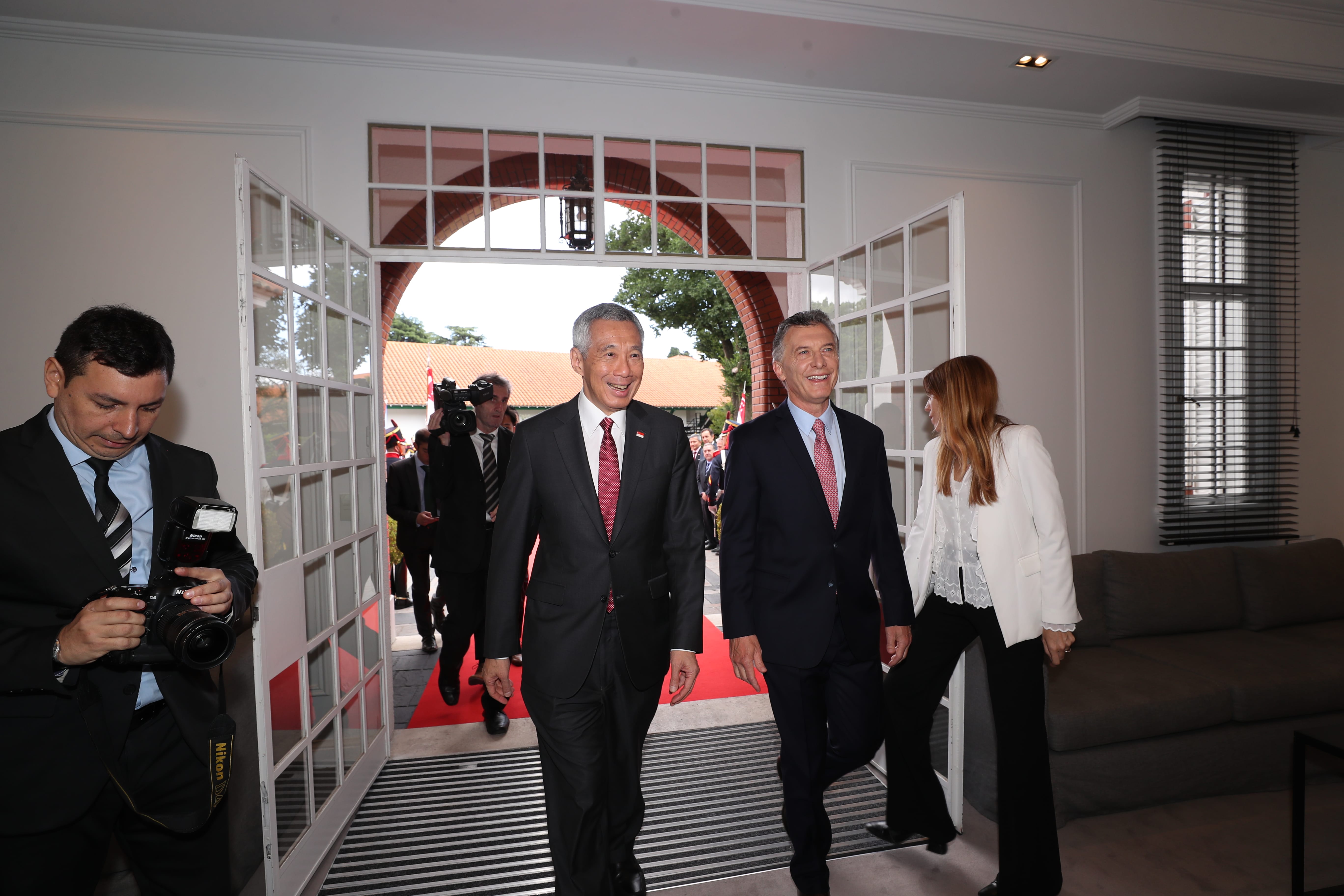 Lee Hsien Loong and Mauricio Macri walk through doorway, followed by people. Nikon camera visible.