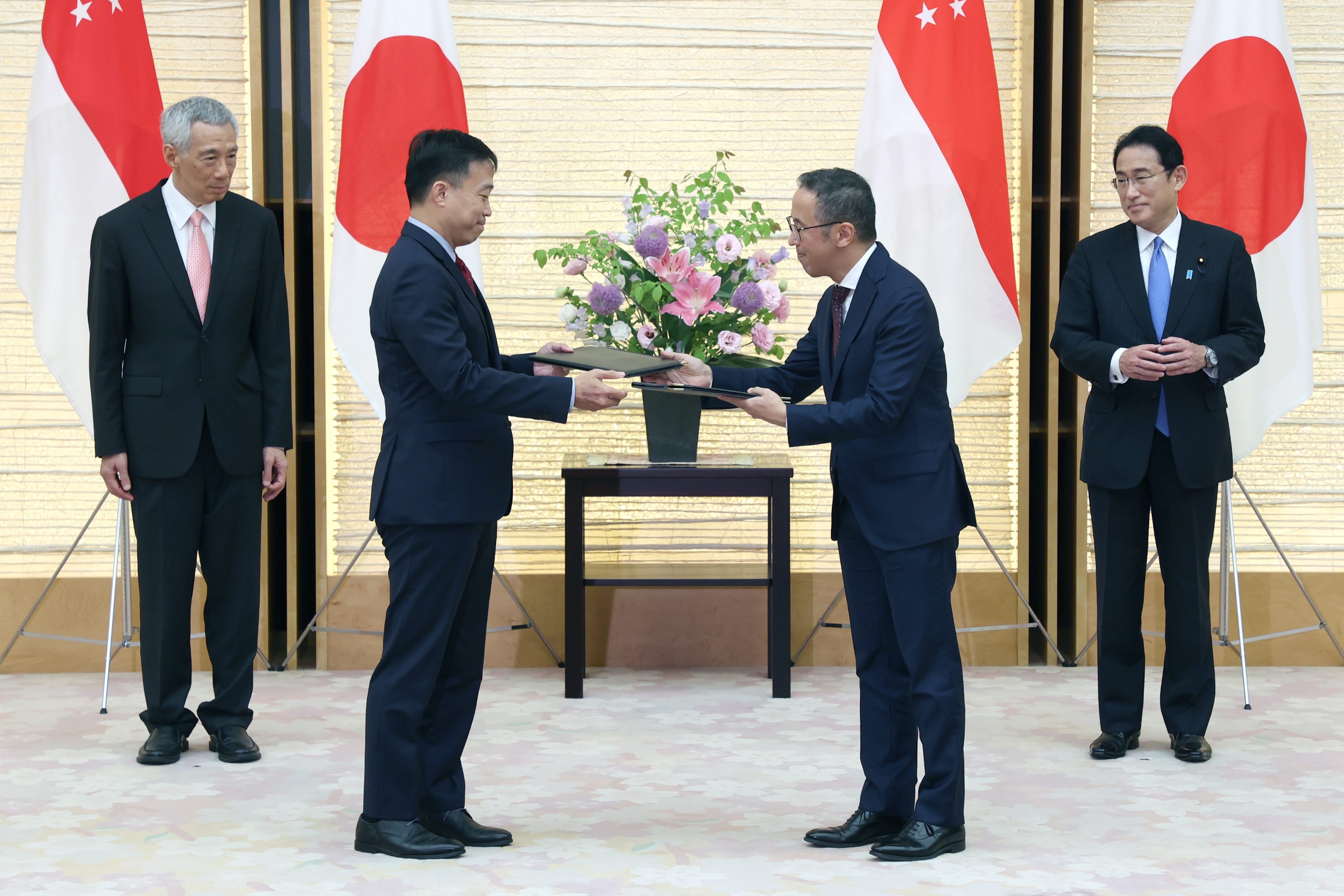 Two men exchanging documents, flags of Japan and Singapore in background, floral arrangement between them.