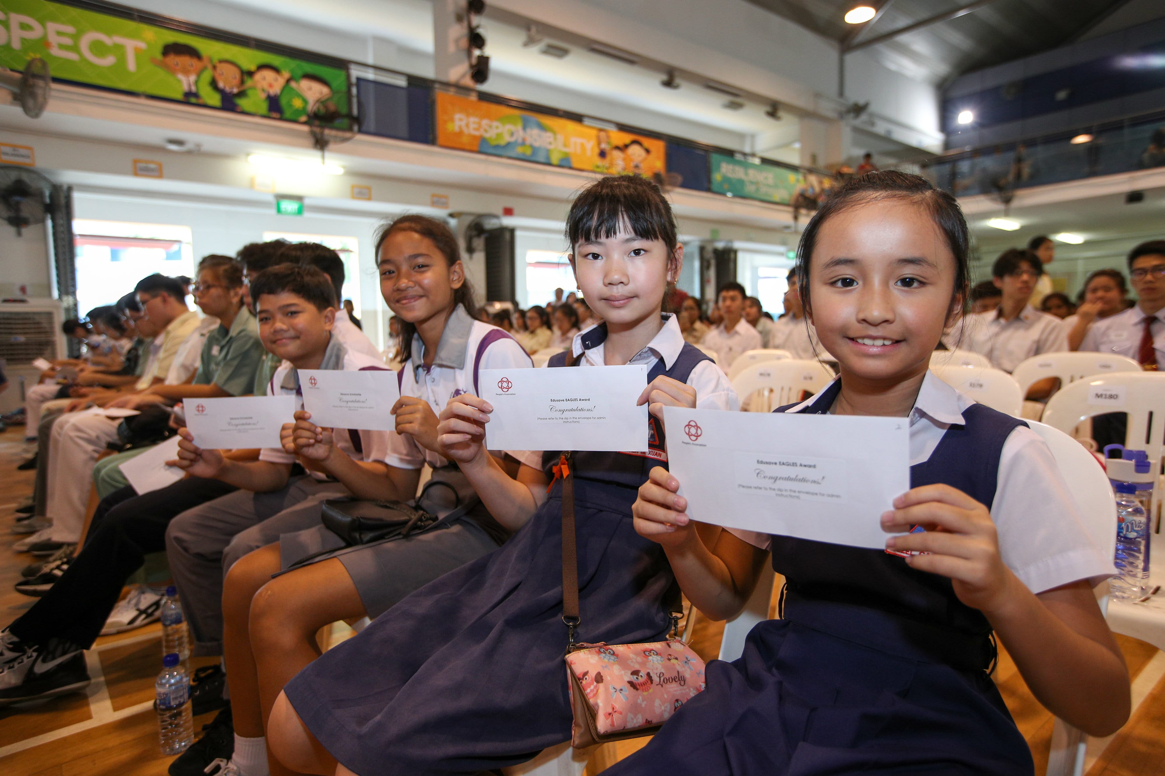 Students in school uniforms hold Edusave EAGLES Award envelopes, sitting in rows at an award ceremony.