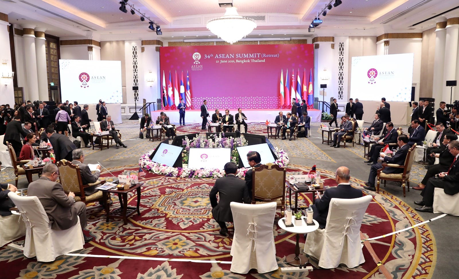 ASEAN summit room with people seated, flags, and screen displaying event details.