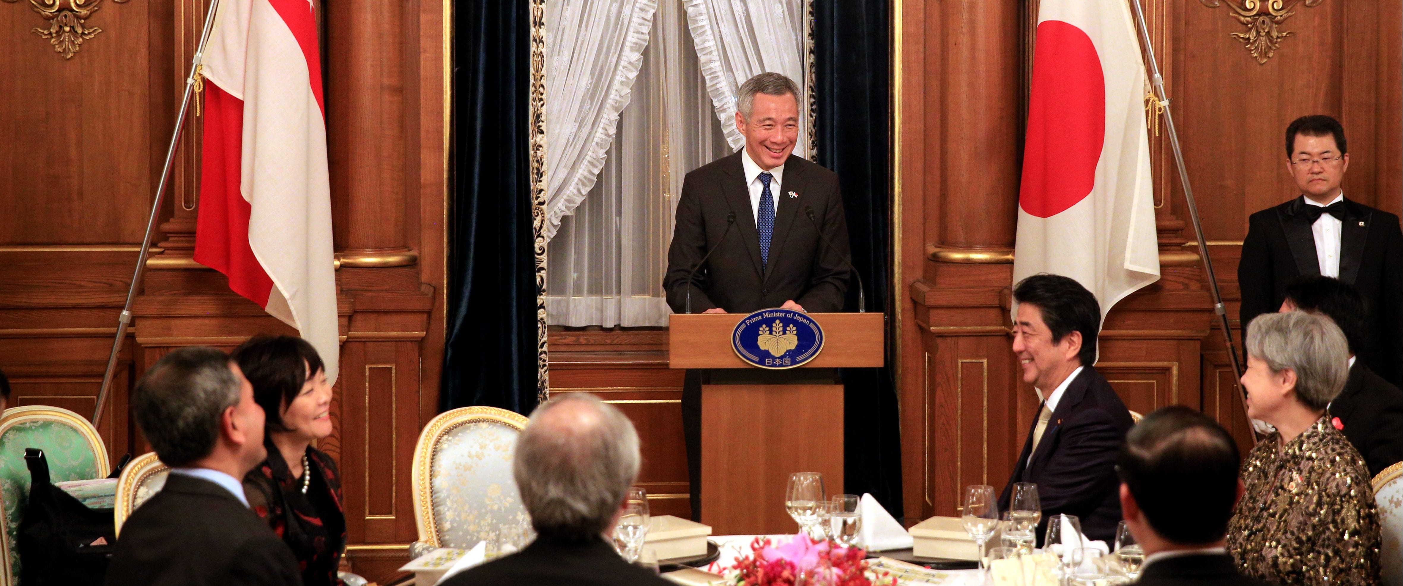 Lee Hsien Loong at podium with Singapore, Japan flags, attended by Shinzo Abe and others at dinner.