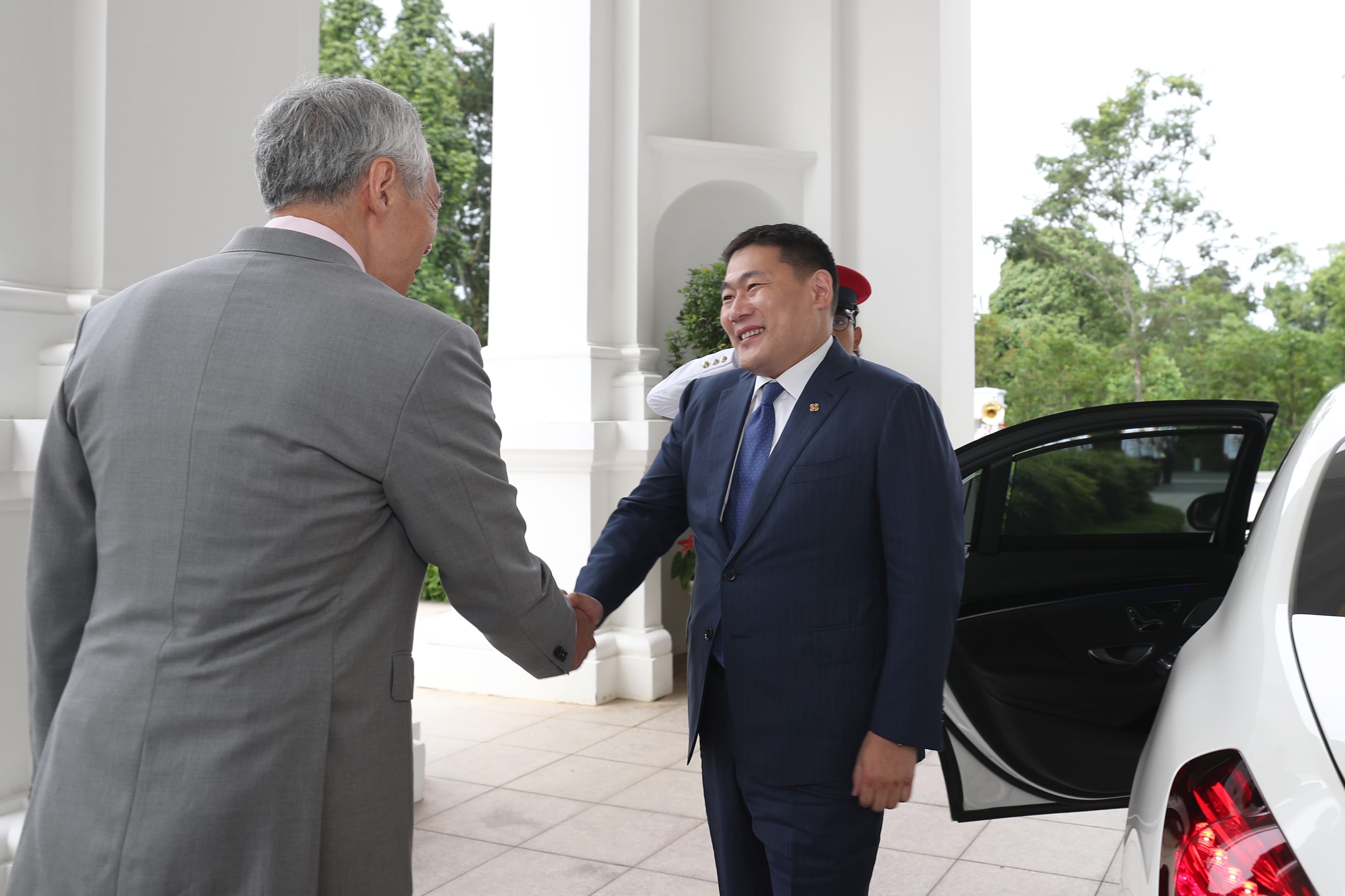 Two men in suits shaking hands, one next to a white car with an open door.