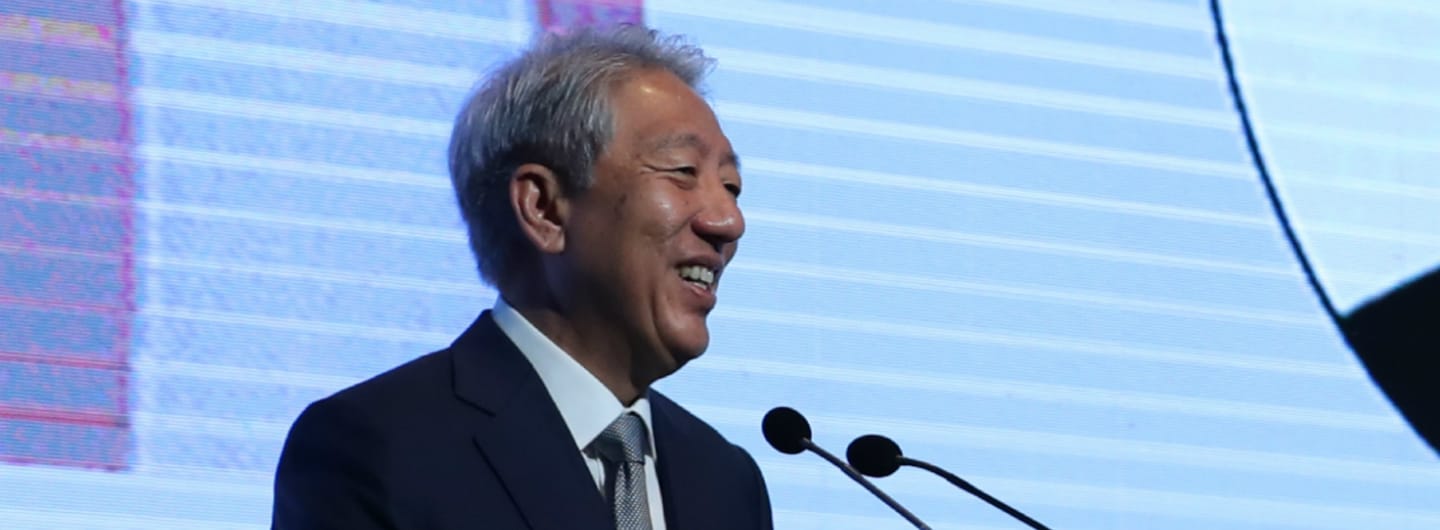 Smiling Asian man in suit speaking at podium with dual microphones, blue patterned backdrop.